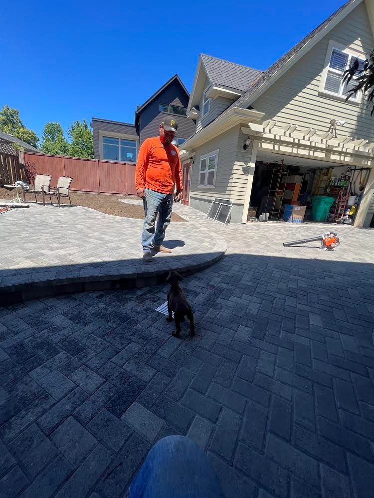 Man in orange shirt stands near small dog on patio with house in background.