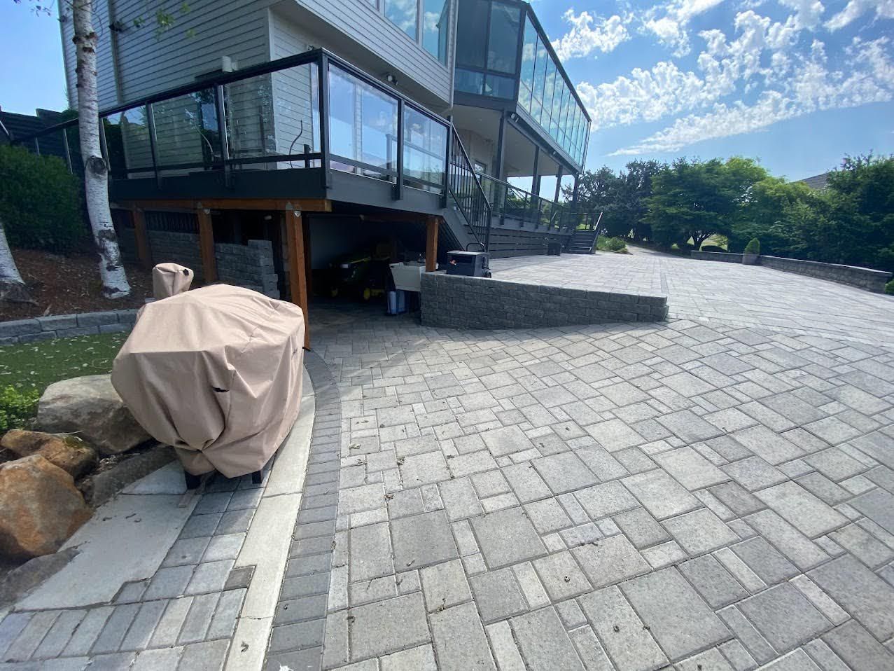 Paver driveway leading to a modern house with a deck. A grill is covered. Sunny day.