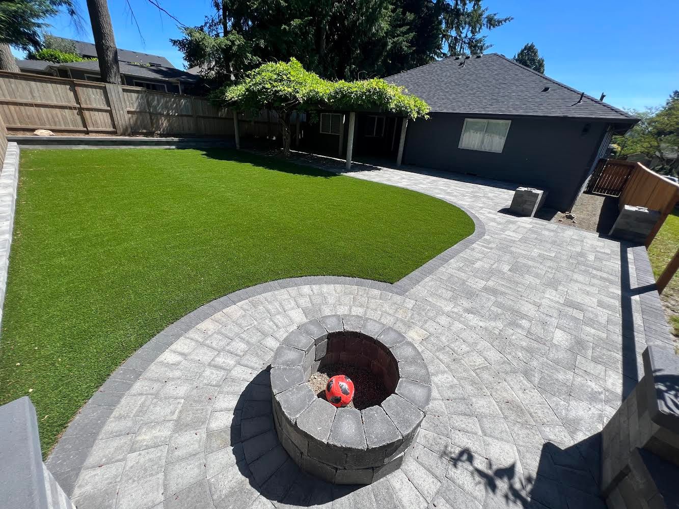 Backyard with fire pit, patio, artificial turf, and a small building with a dark roof under a blue sky.