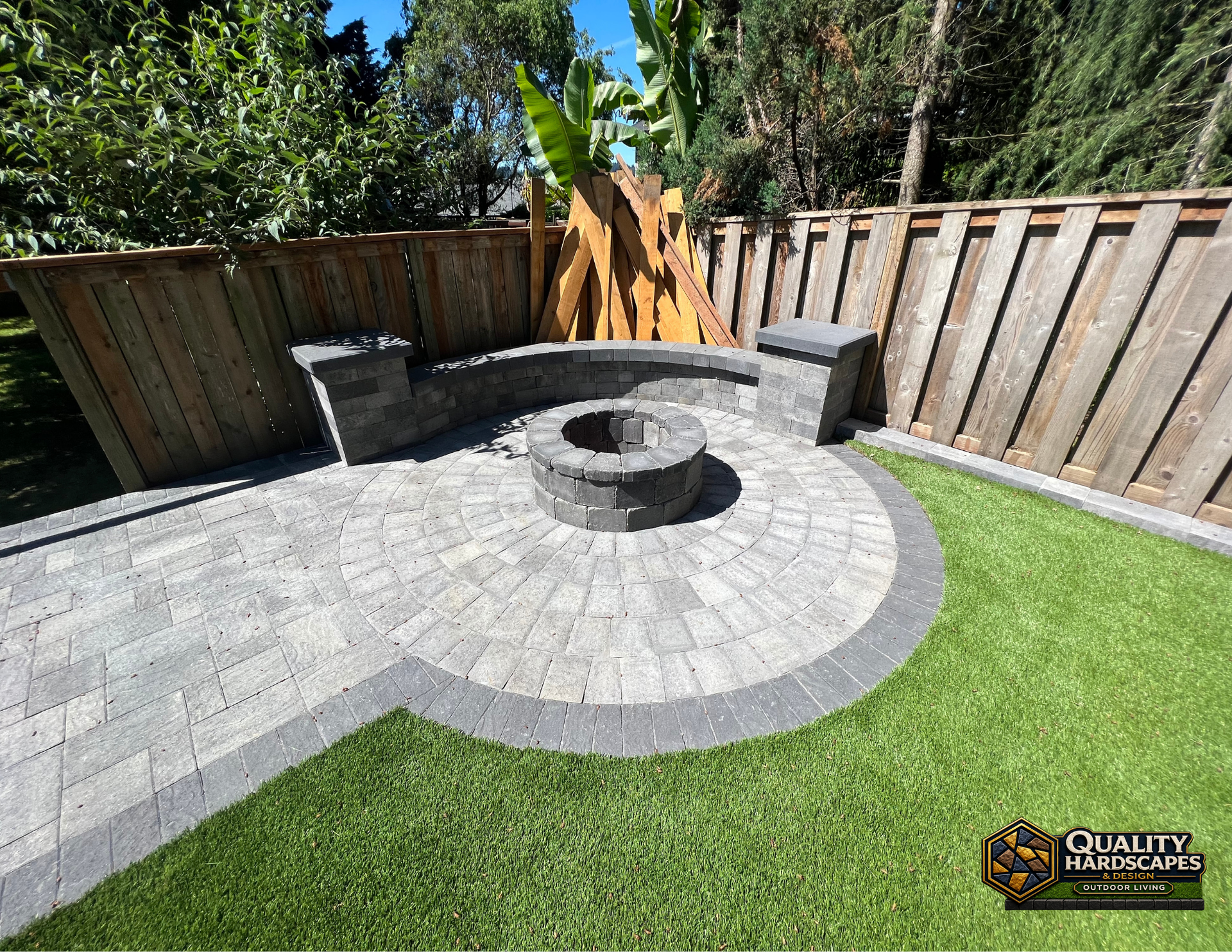Backyard patio with curved gray pavers, circular fire pit, and wooden fence beside green lawn