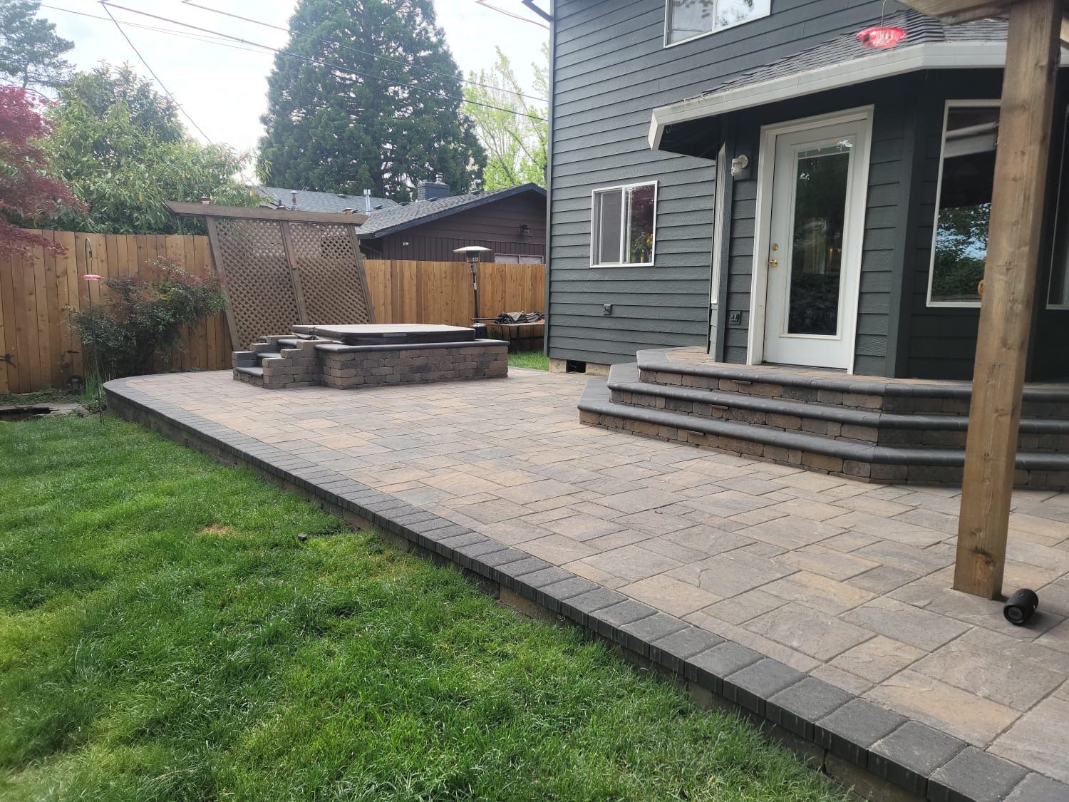 Patio with pavers and steps, hot tub area, and a gray house in the background.