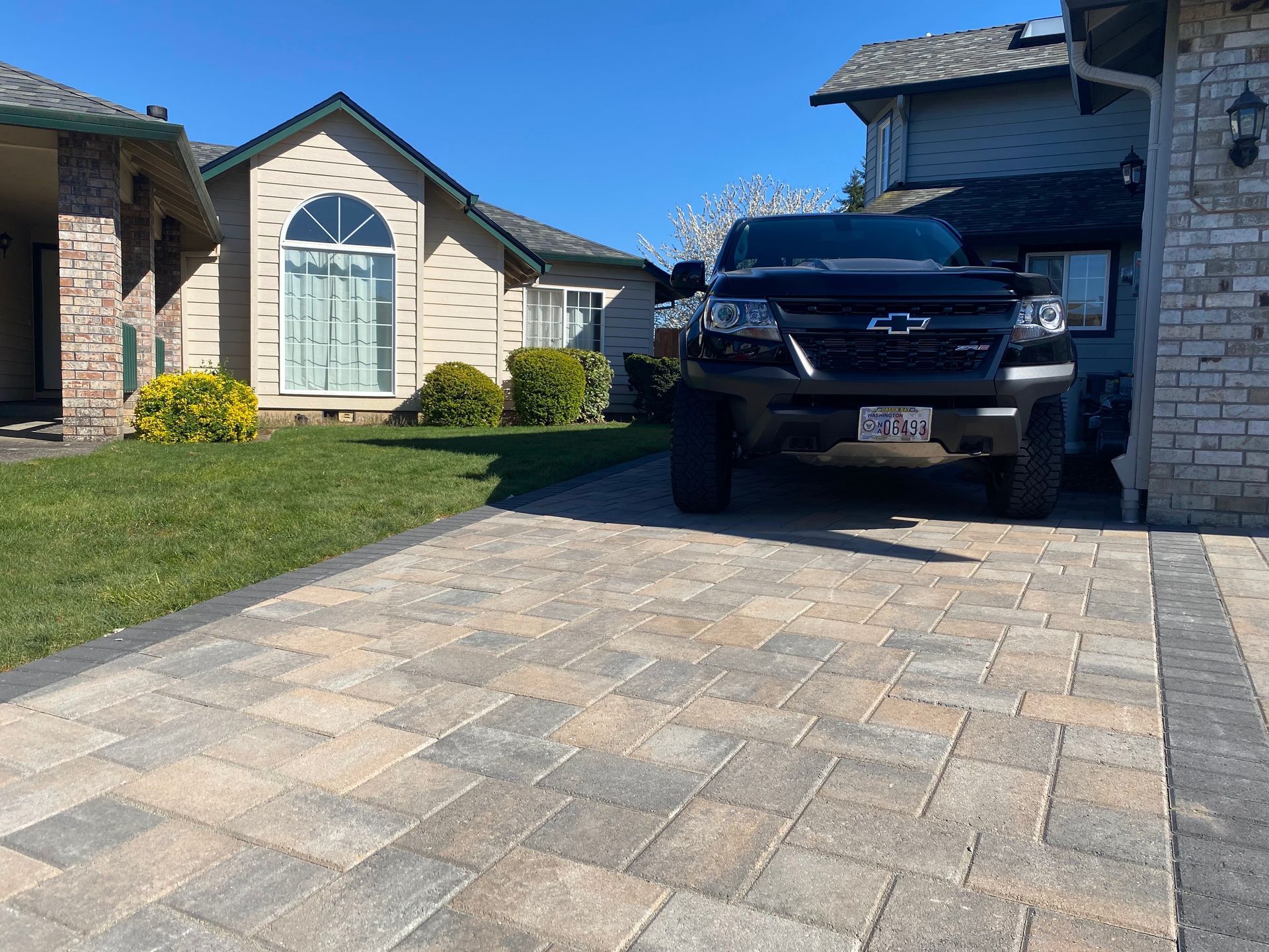 Black pickup truck parked in front of a house on a brick driveway, with grass and bushes on a sunny day.