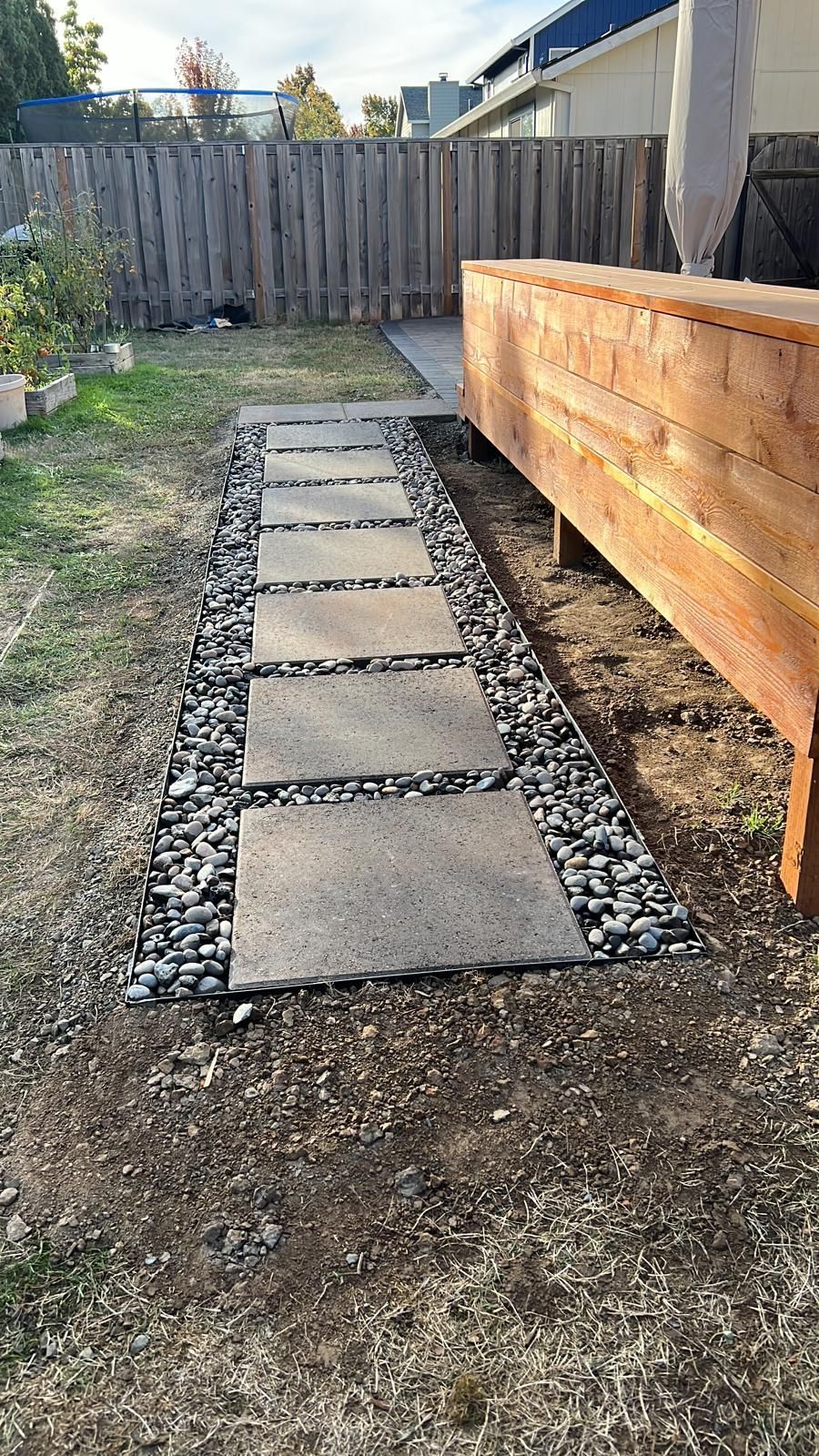 Stone path with stepping stones through a backyard, bordered by black pebbles, next to a wooden deck.