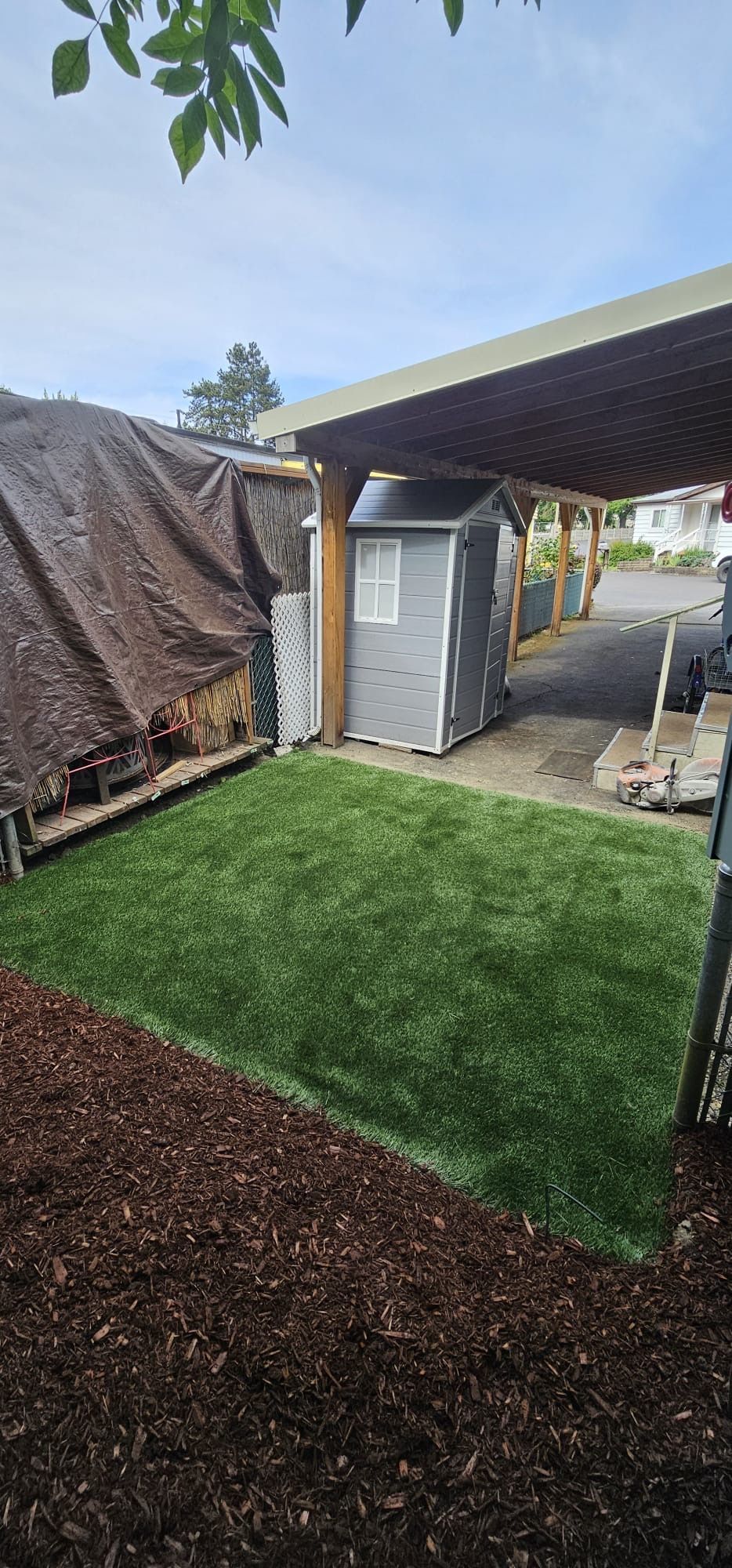 A small gray shed in a yard, bordered by fresh mulch, next to green artificial turf.