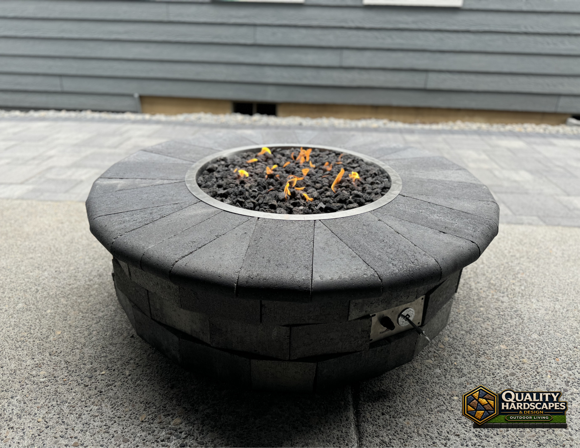 Round gray outdoor fire pit with embers in a paved patio area near a house siding