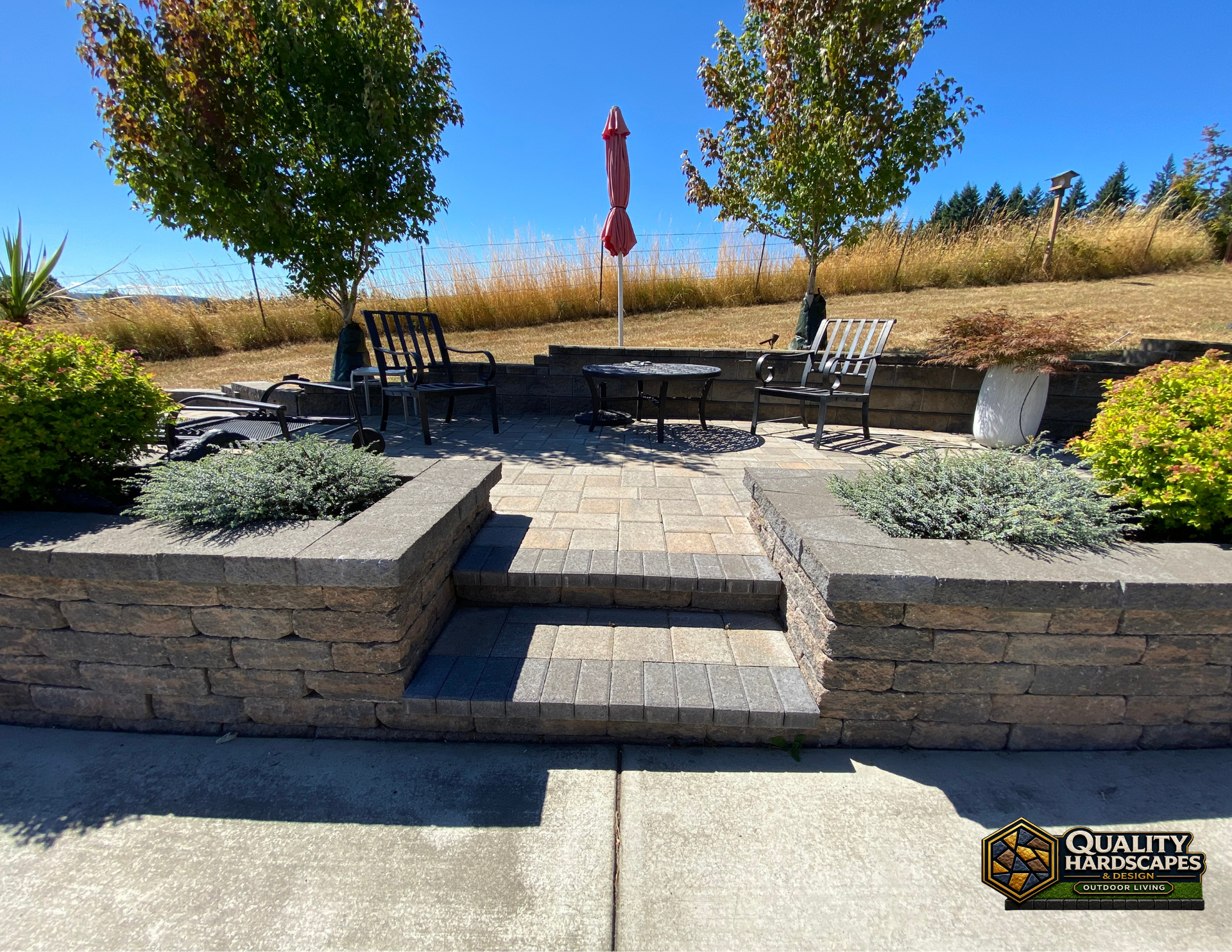 Backyard patio with stone steps, black outdoor chairs, red umbrella, and landscaped planters.