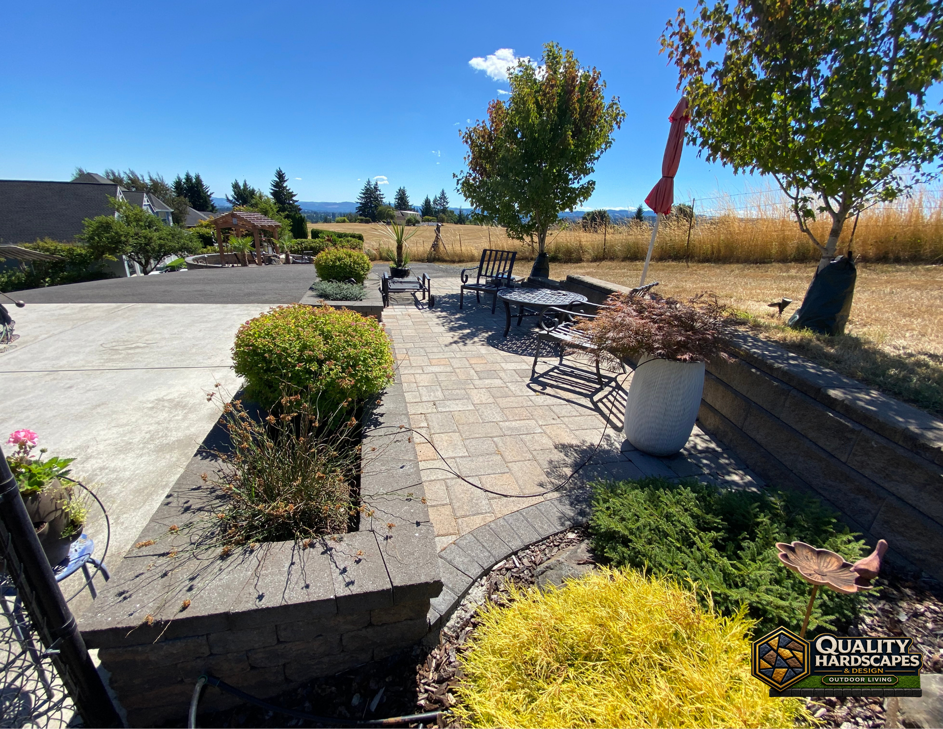 Patio with tables, potted plants, and stone walkway beside a field under a bright blue sky