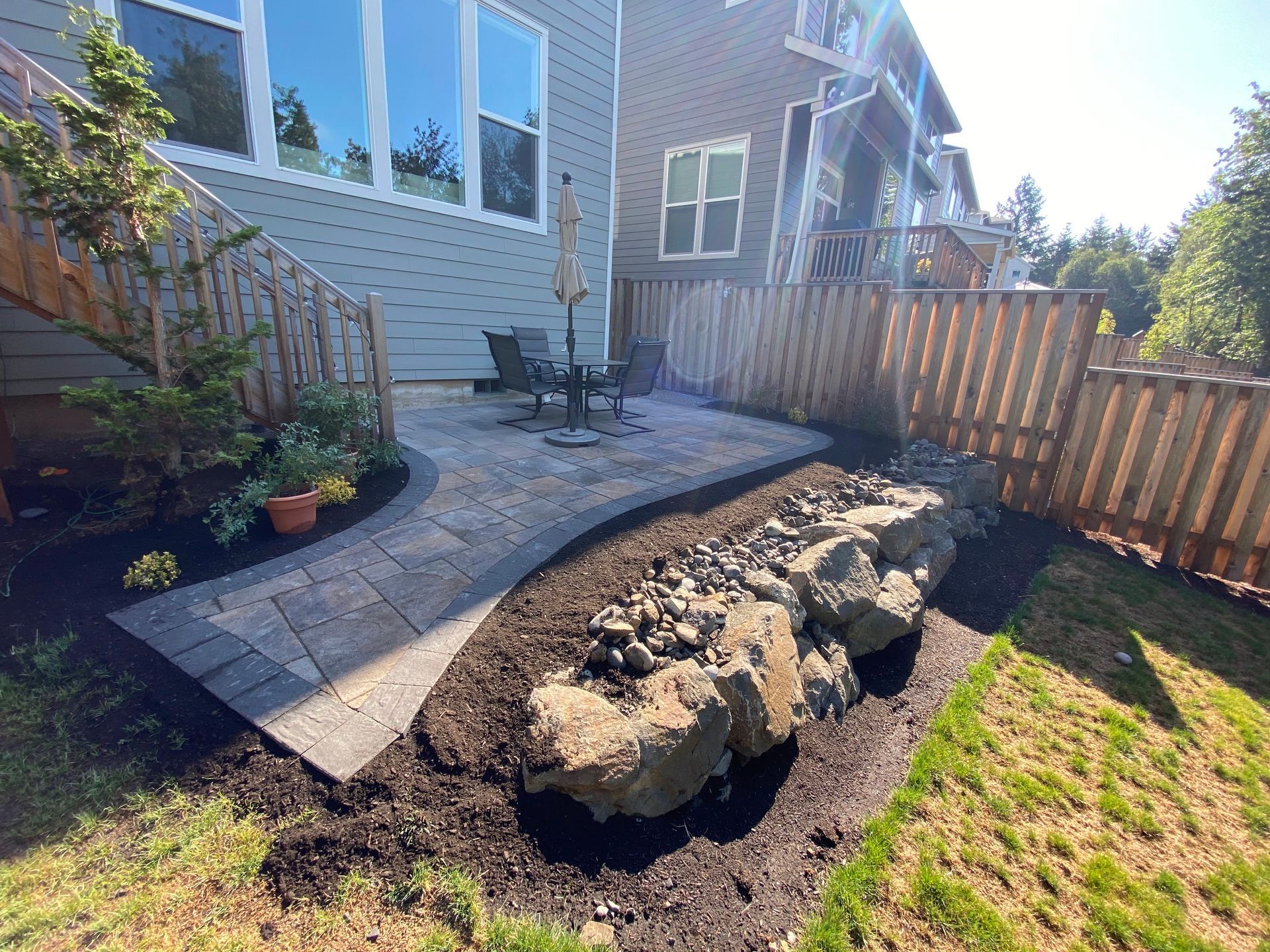 Backyard patio with stone walkway, landscaping, rock feature, and wooden fence.