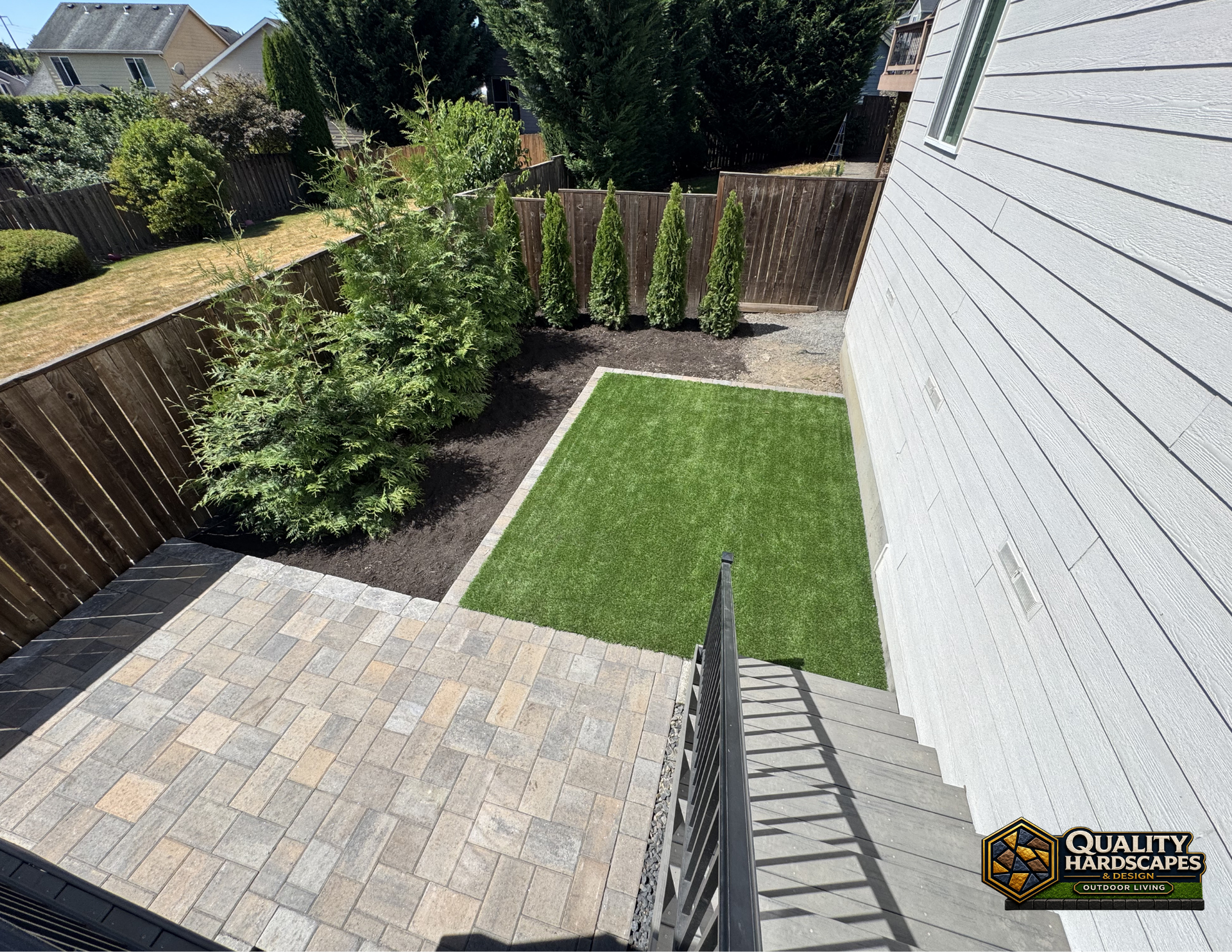 Backyard with gray paver patio, small lawn, and landscaped shrubs beside a white house