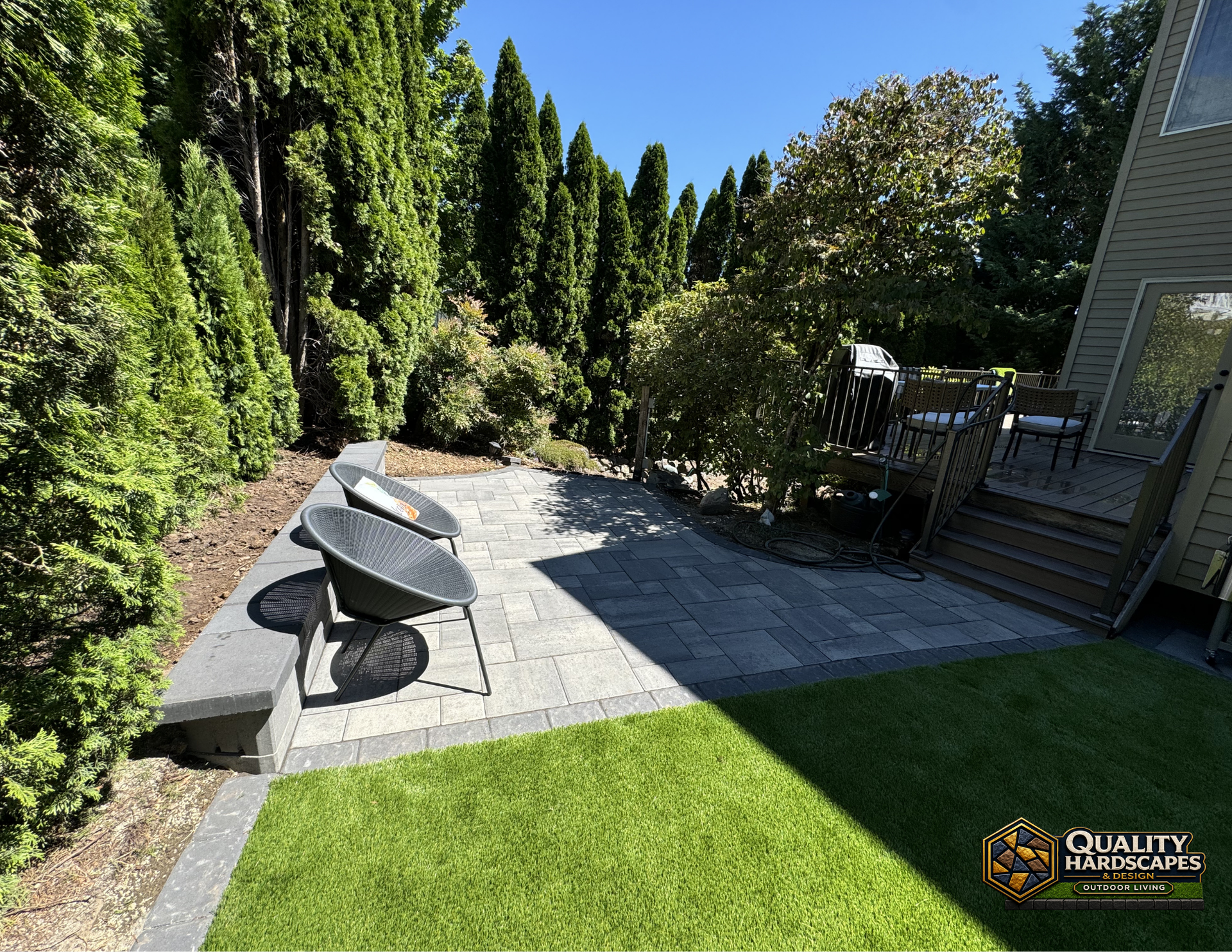 Backyard patio with stone pavers, two lounge chairs, and a lush green lawn beside a house.