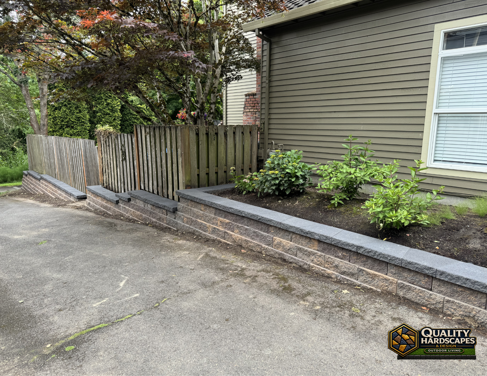 Side yard with a wooden fence, raised garden bed, and shrubs beside a house and driveway.