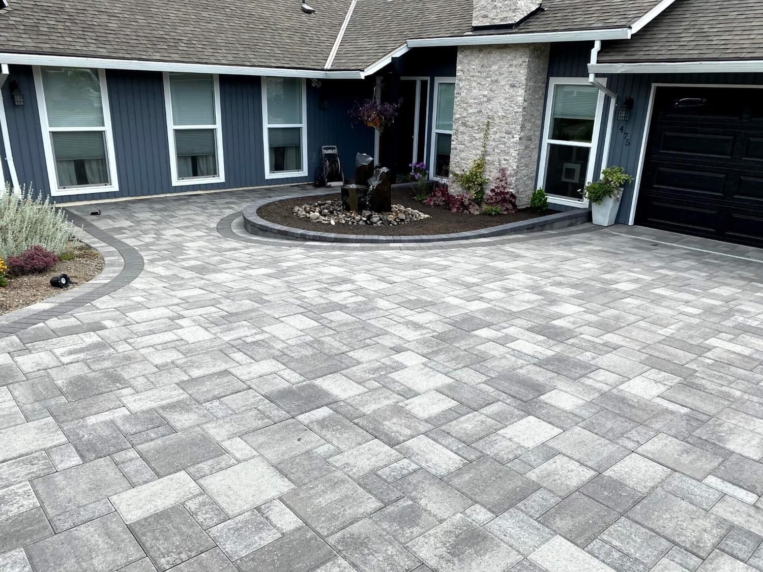 Gray paver driveway leads to a blue house with windows, a brick pillar, and a black garage door.