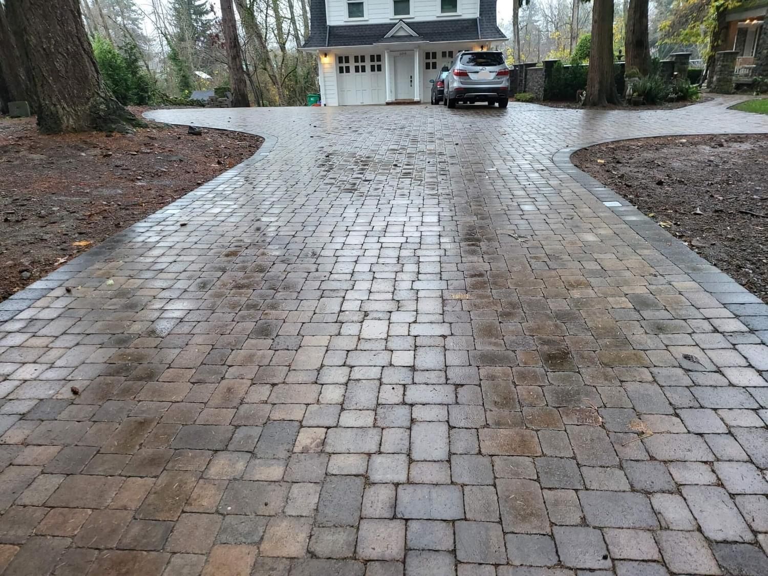 Brick driveway leading to a white house with a car parked in front of a garage. Trees frame the scene.