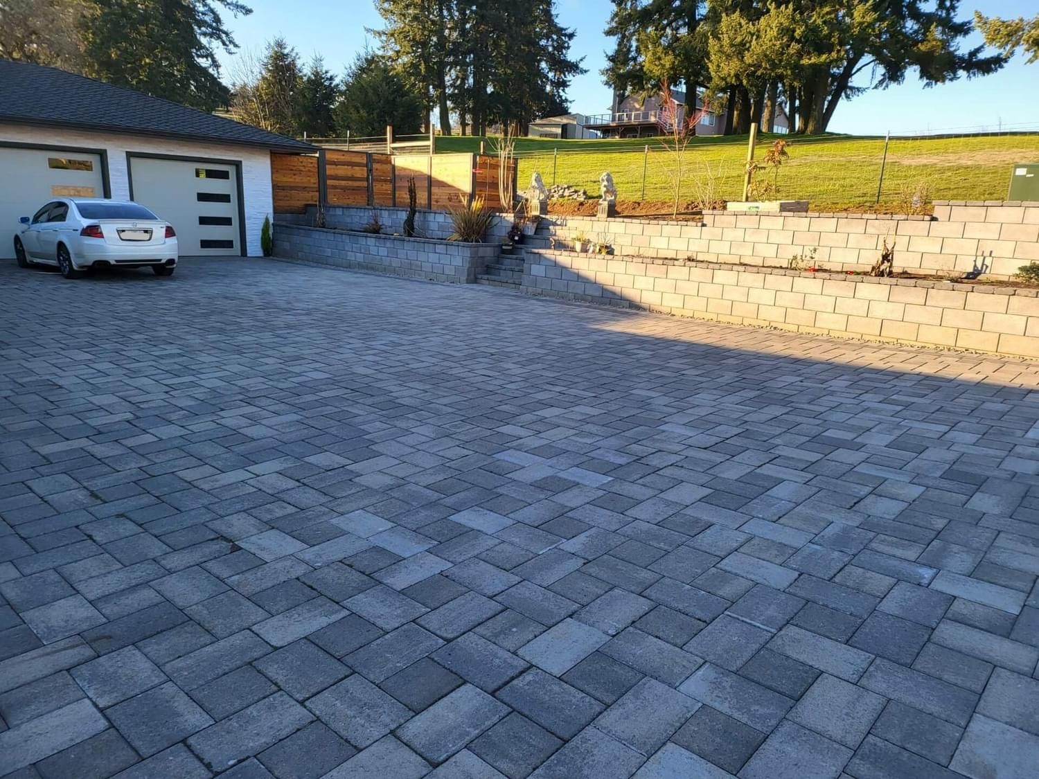 Driveway paved with gray bricks, leading to a garage and a hillside with trees and a building in the background.