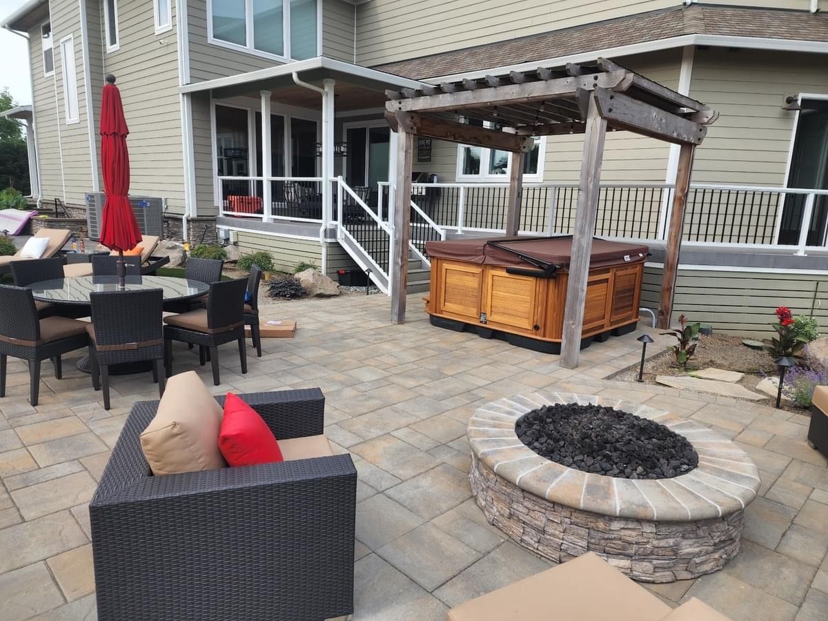 Outdoor patio with hot tub under a wooden pergola, fire pit, and seating. House in the background.