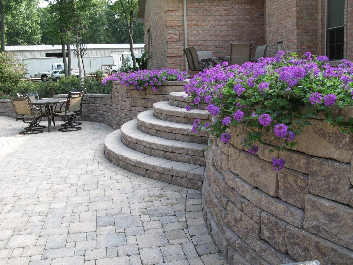 Curved brick patio with steps and retaining walls, purple flowers, and outdoor seating.