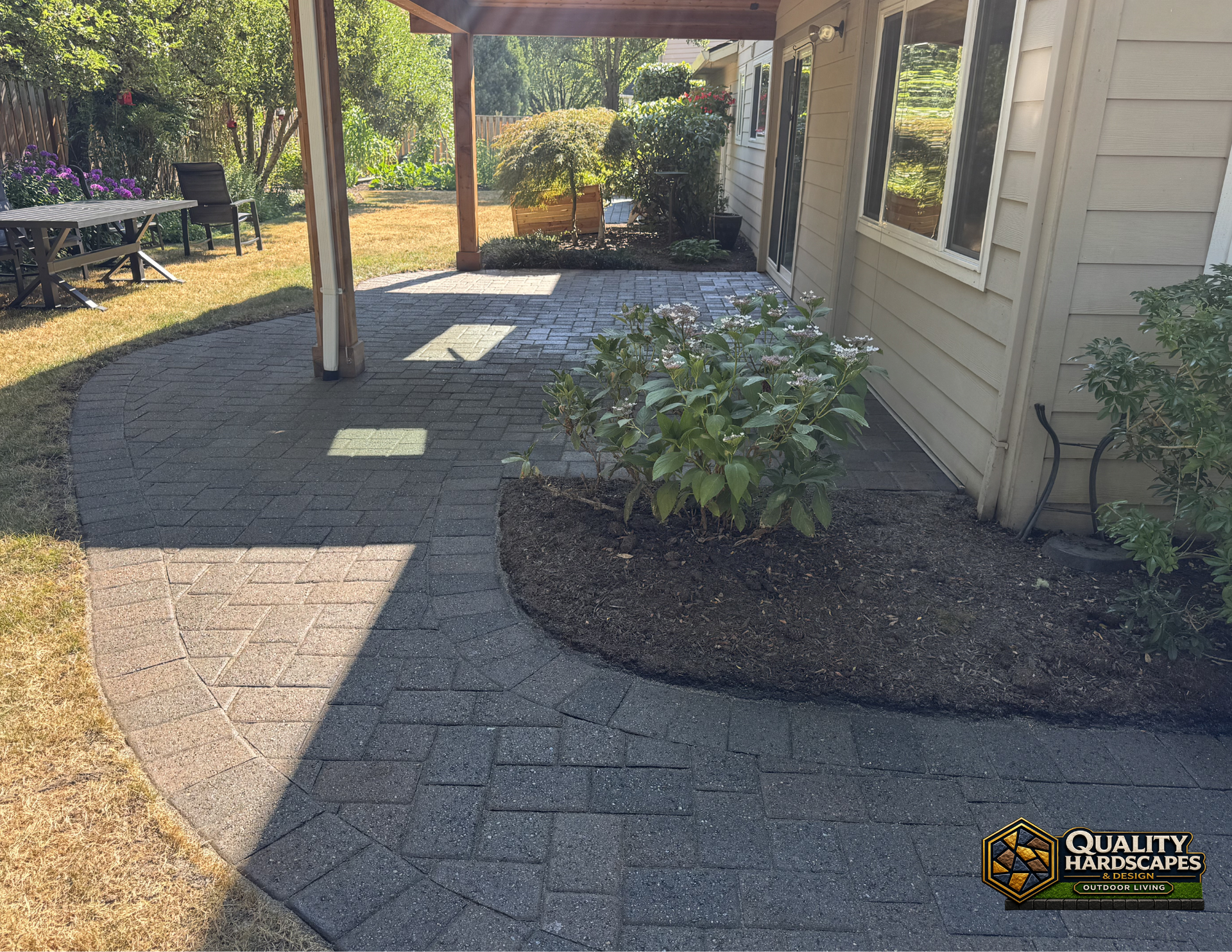Curved paver walkway beside a house and covered patio, bordered by mulch and shrubs.