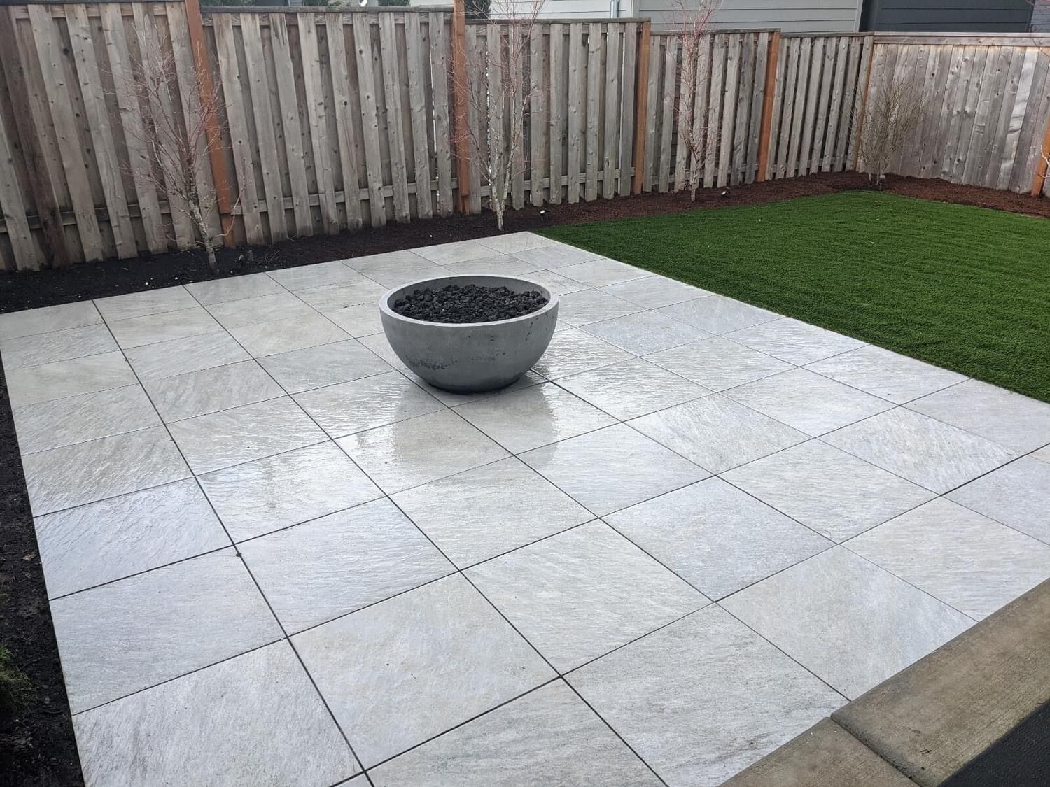 A concrete patio with a fire pit, bordered by a wooden fence and artificial grass.