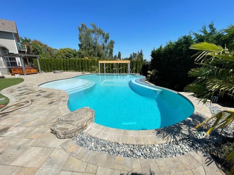 A large, curved swimming pool with blue water. Stone patio surrounds it, with a pergola and greenery in the background.