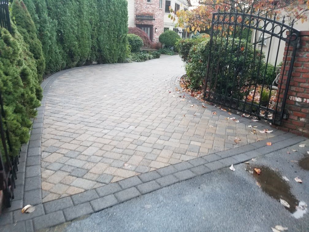 Brick driveway curves toward a house, with hedges and a wrought iron gate.