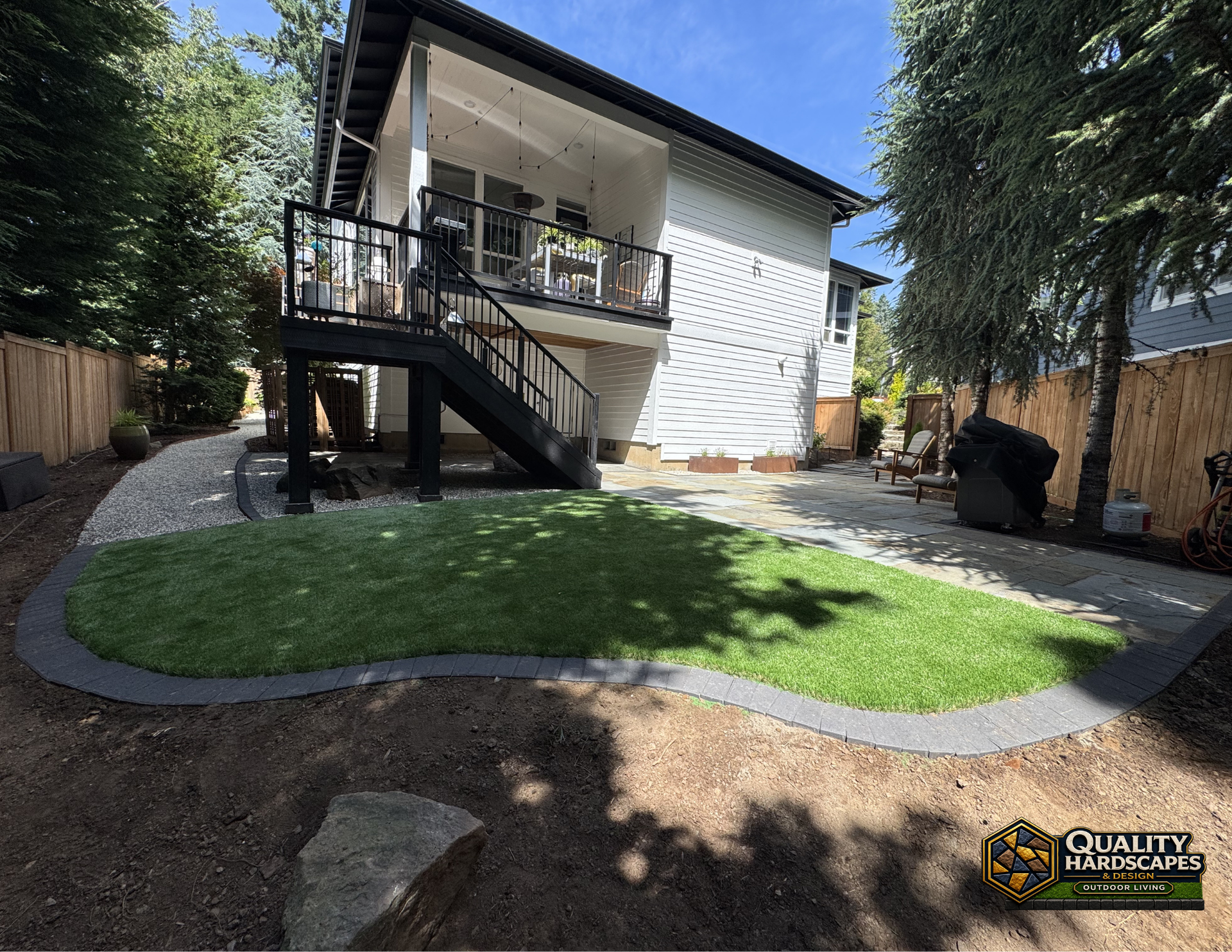 Backyard patio with raised deck, stairs, and green lawn beside a white house