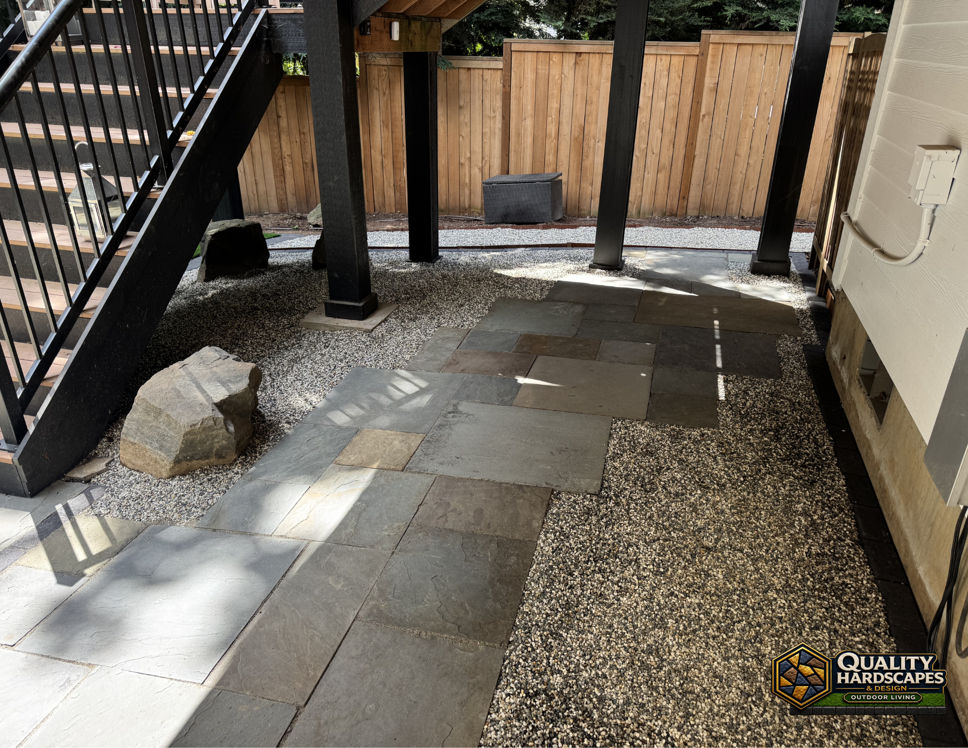 Outdoor patio with stone pavers, gravel, wooden fence, and black metal stairs in partial shade