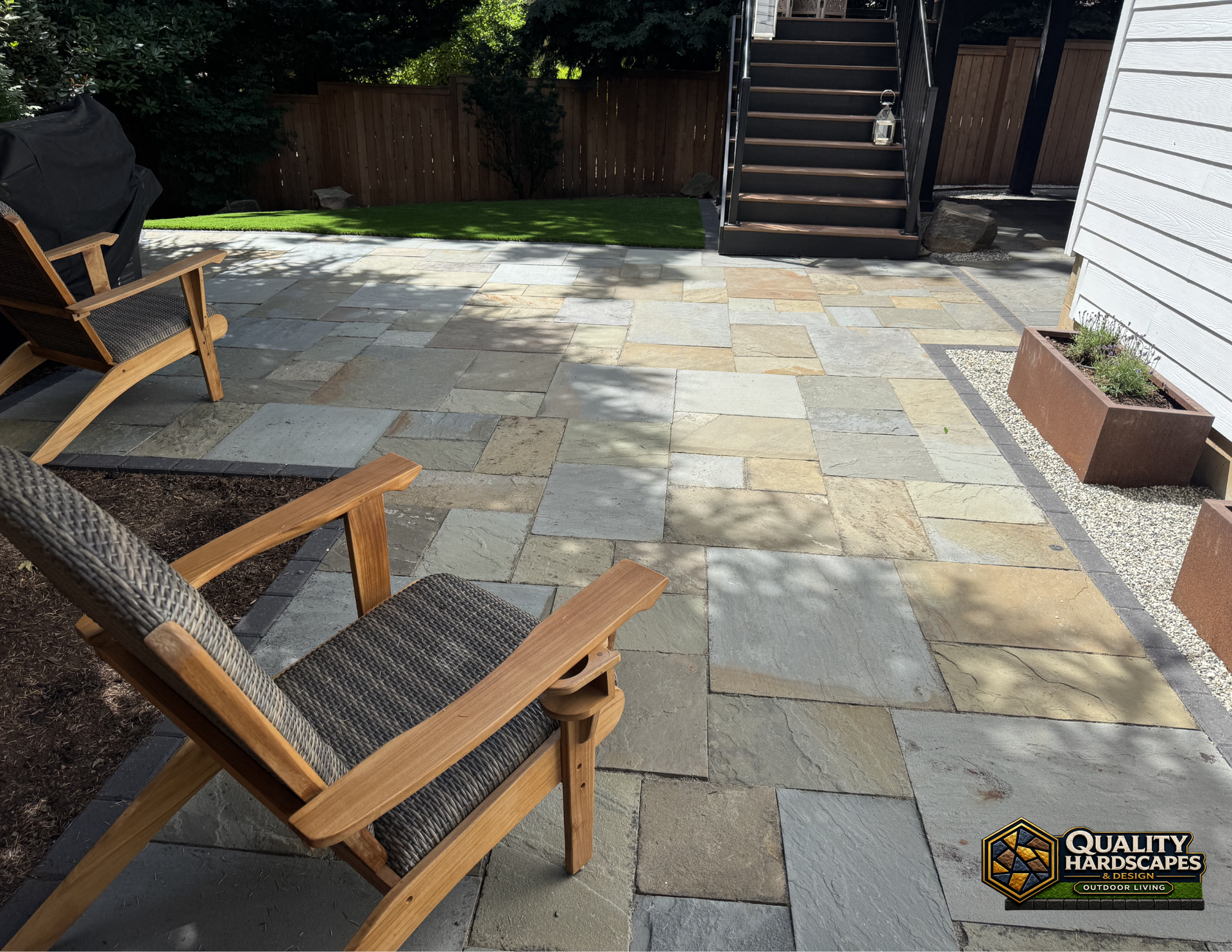 Patio with light stone pavers, wooden chairs, stairs, and a planter beside a house.