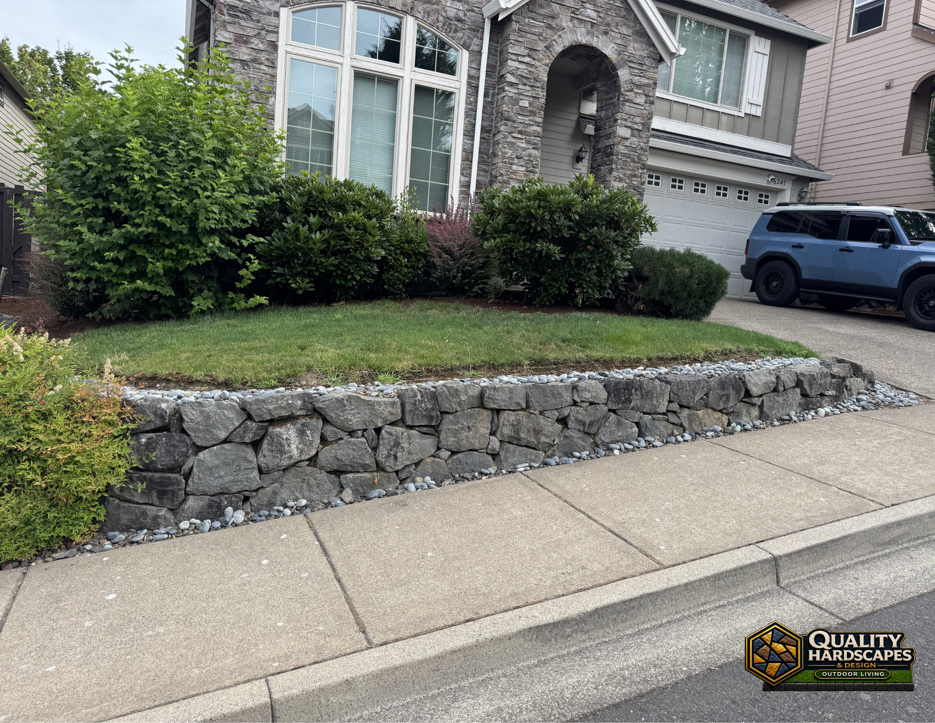 Stone retaining wall and shrubs in front of a suburban house with a driveway and parked SUV.