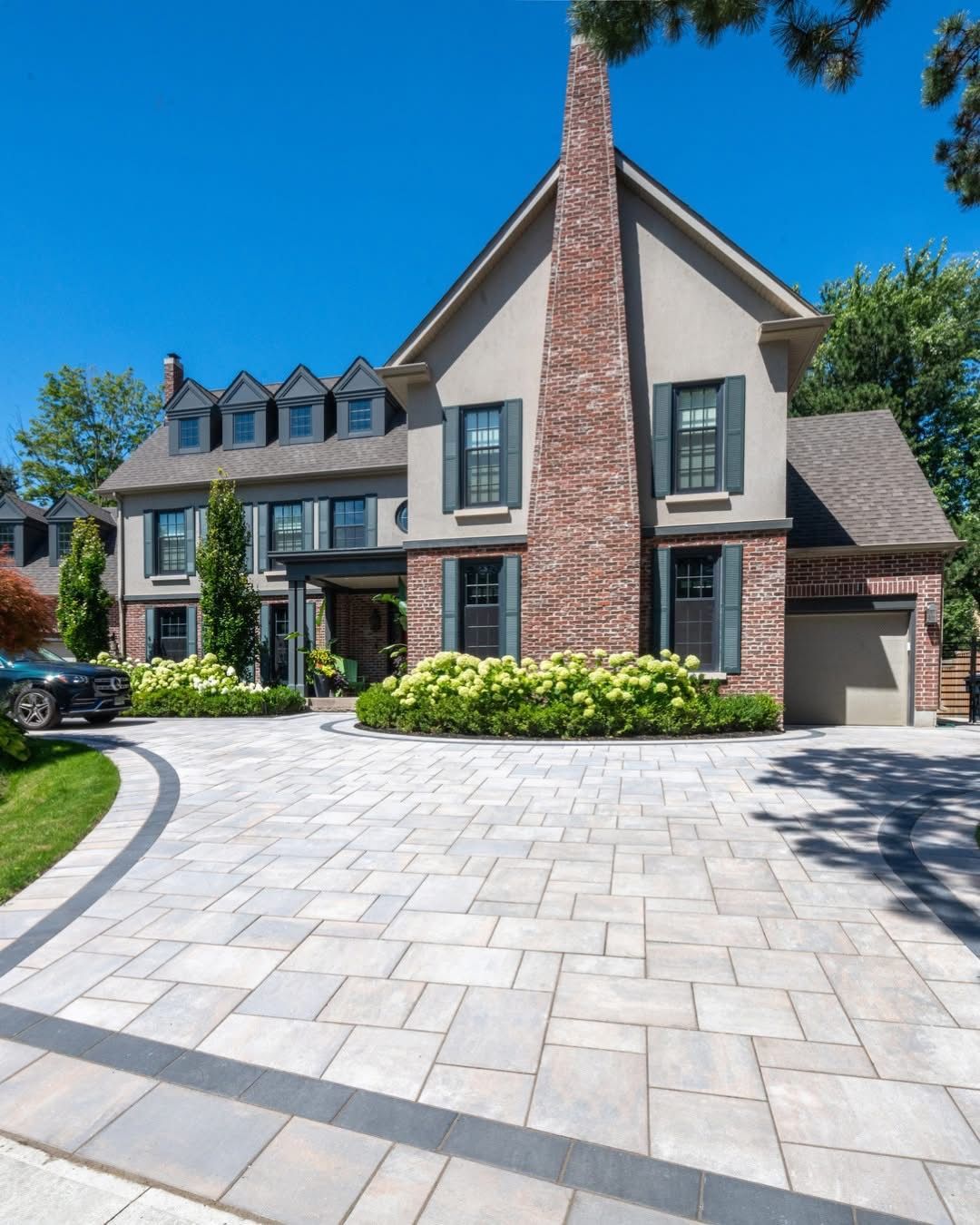Elegant two-story house with brick chimney, gray facade, and patterned driveway on a sunny day.