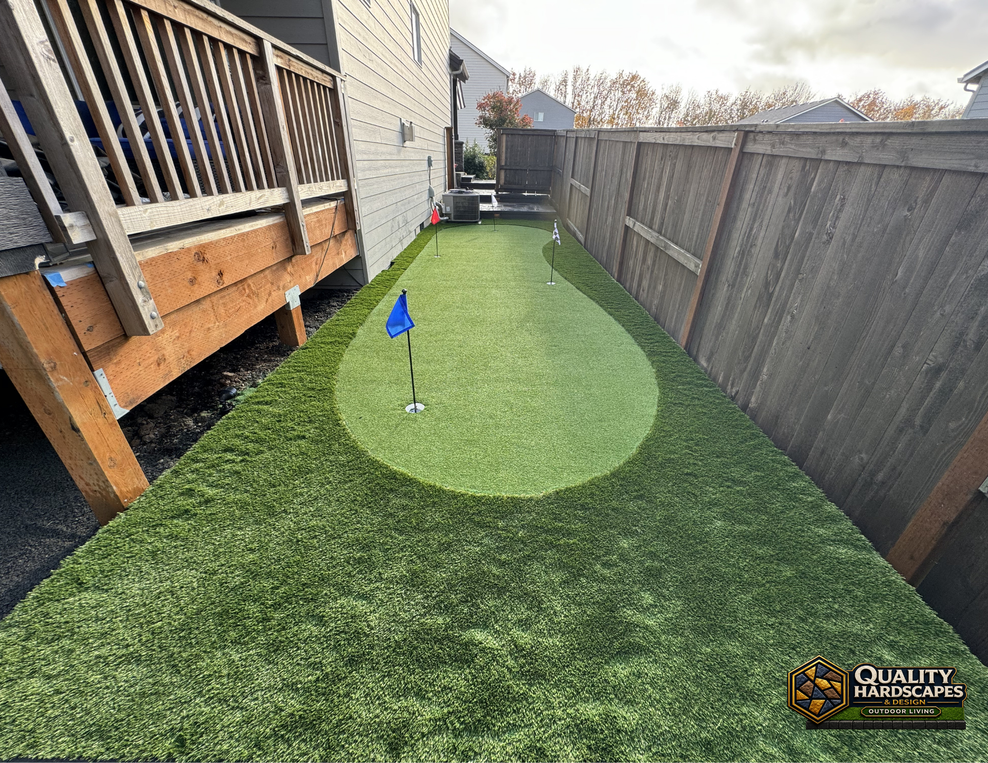 Backyard putting green with a golf hole, blue flag, and wooden deck beside a fence