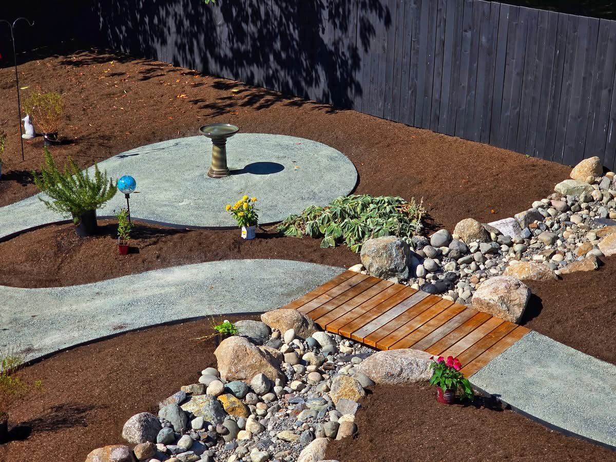 Gravel paths and wooden bridge in a landscaped yard with a birdbath and rock-lined stream.