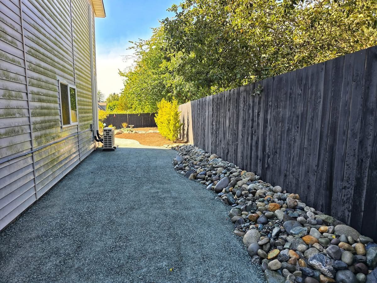 Side yard with gravel, rocks, wooden fence, and building exterior. Sunny day with trees in the background.