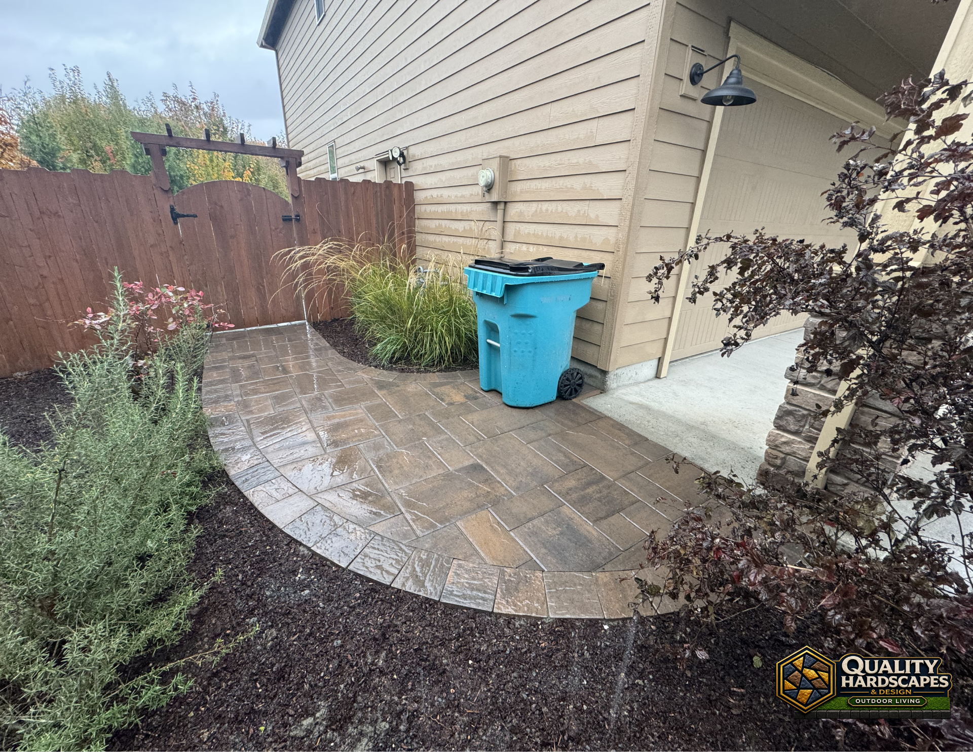 Curved stone patio beside beige house with blue recycling bin and landscaped garden border