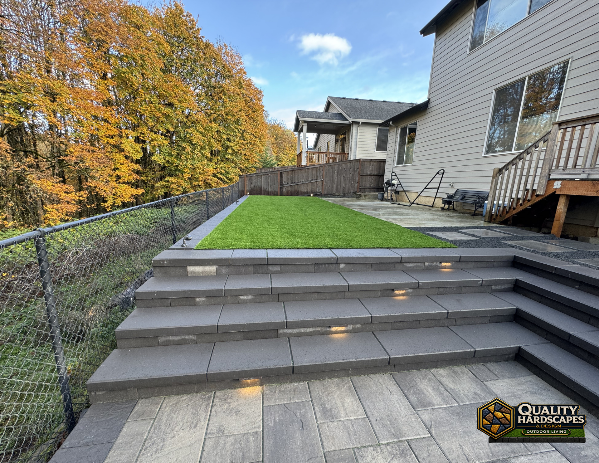 Backyard stone steps with gray pavers, green lawn, and a house deck beside a chain-link fence.