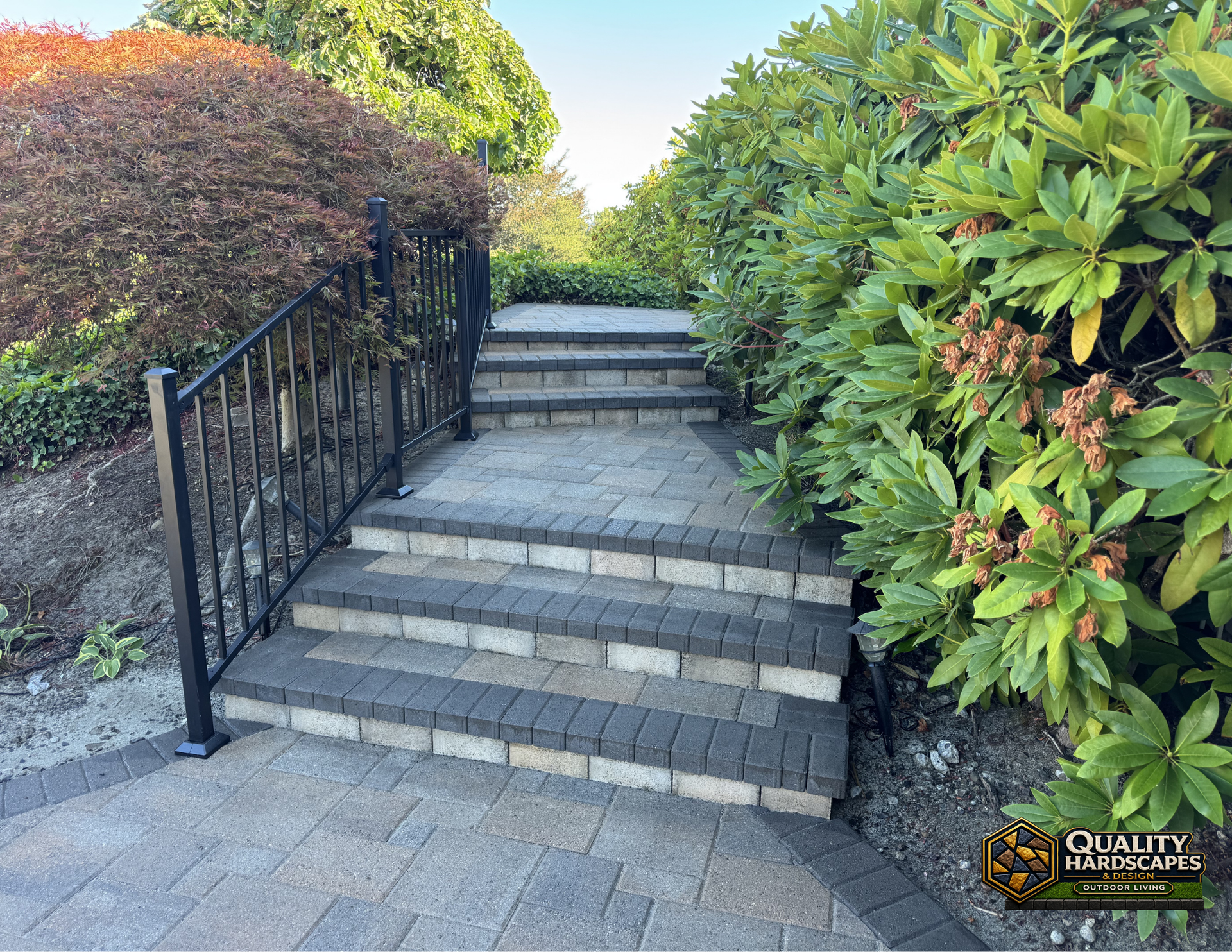 Stone steps with a black handrail between green hedges and shrubs in a landscaped garden.