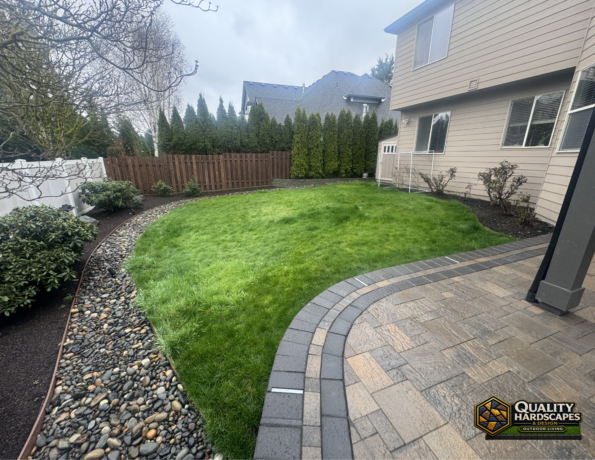 Curved paver patio beside a green lawn and landscaped backyard with shrubs and wooden fence