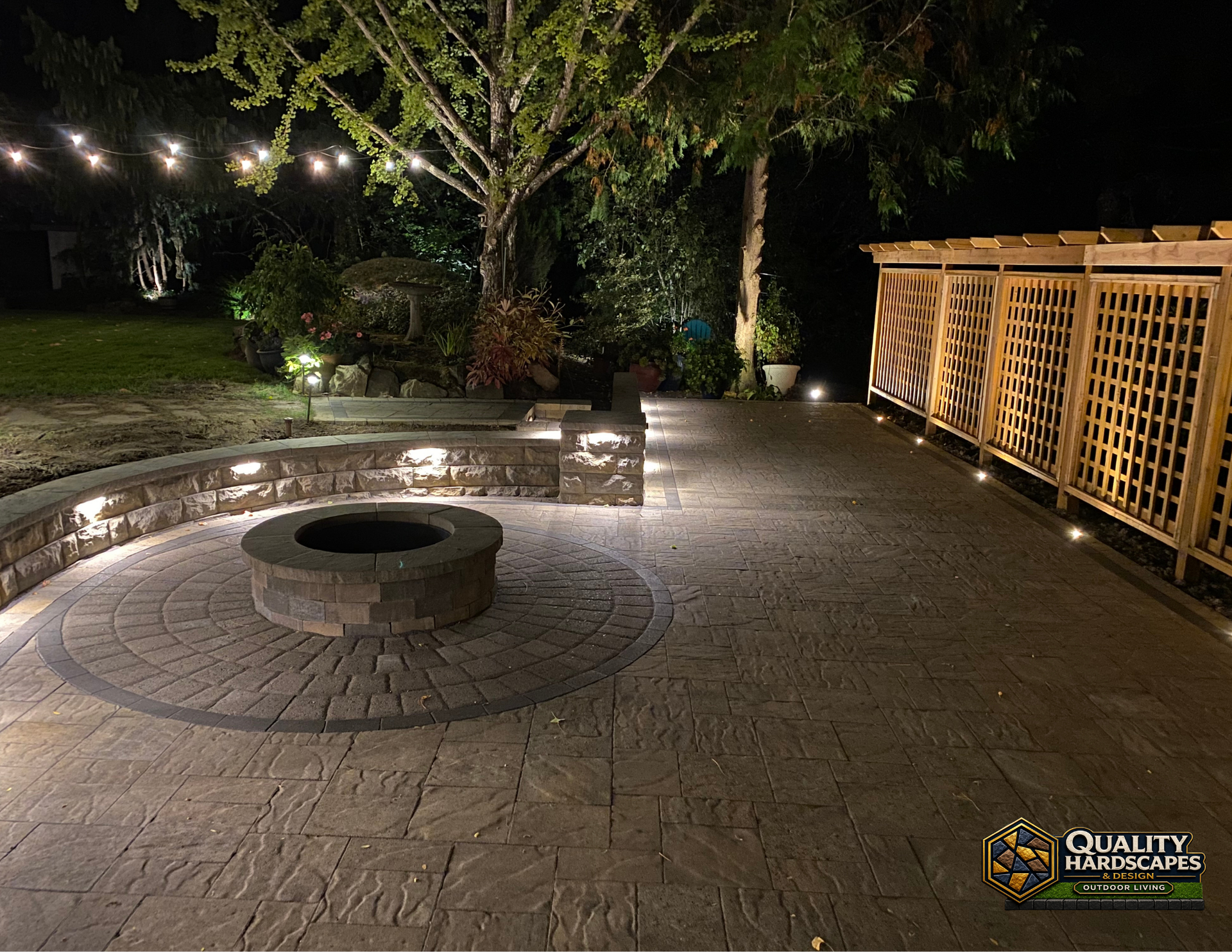 Nighttime patio with stone fire pit, string lights, and a wooden lattice fence beside trees.