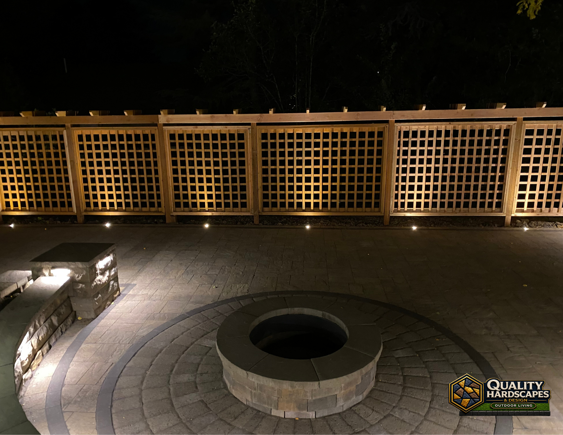 Nighttime patio with a circular fire pit and illuminated lattice fence panels behind it.