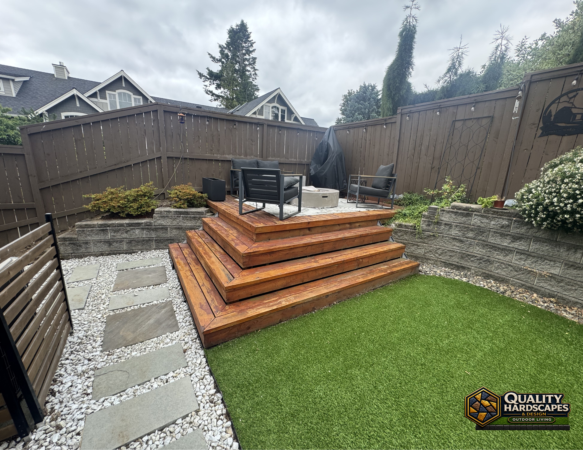 Wooden stepped patio deck with outdoor chairs in a fenced backyard, bordered by stone pavers and grass.
