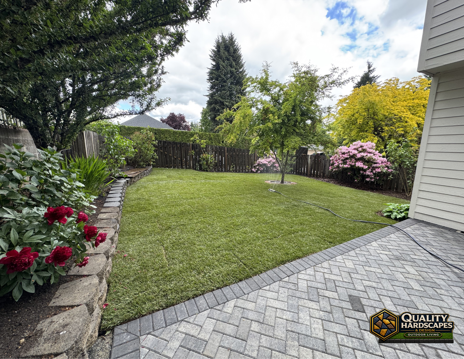 Backyard with green lawn, stone patio, trees, and flowering shrubs beside a house