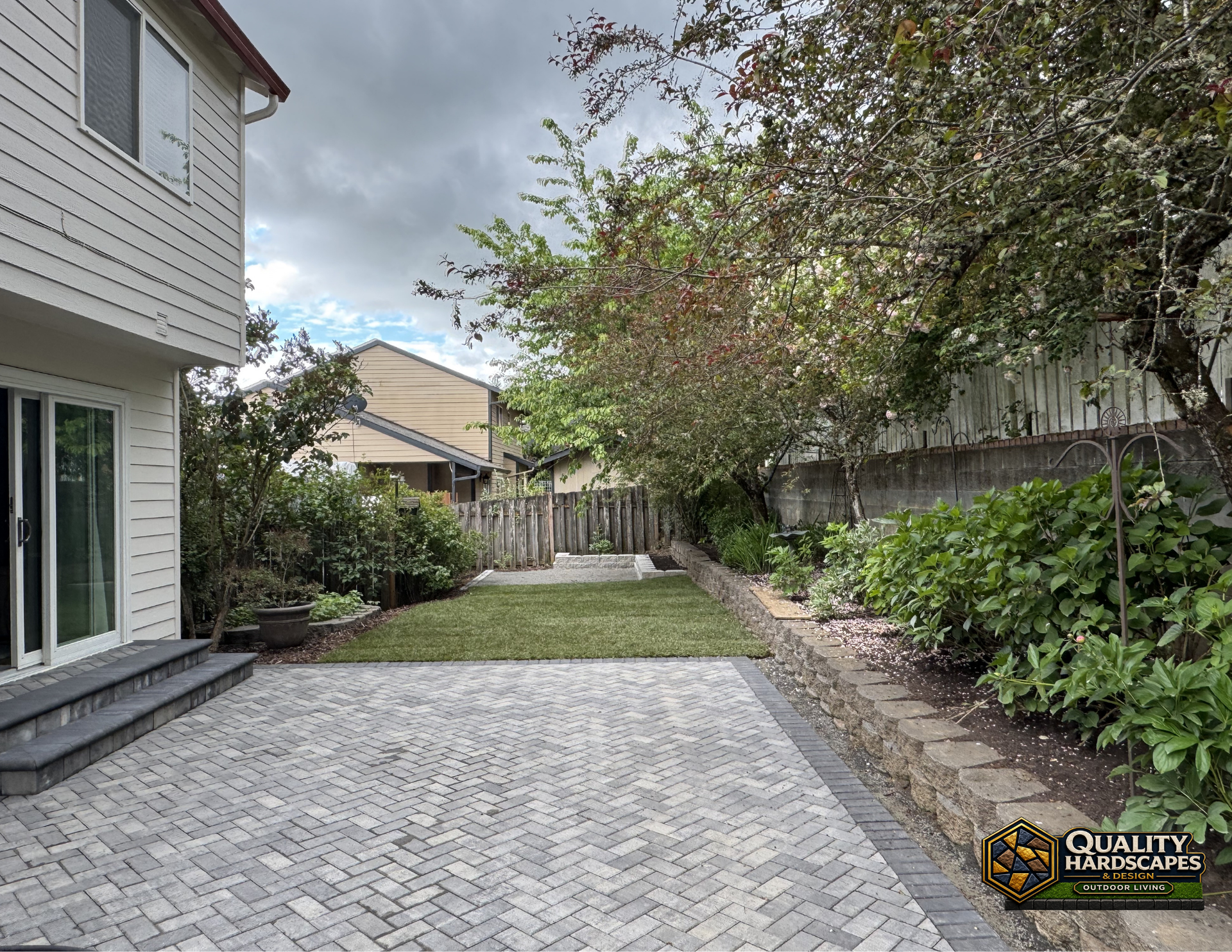 Backyard patio with gray pavers, lawn, shrubs, and a wooden fence beside a house