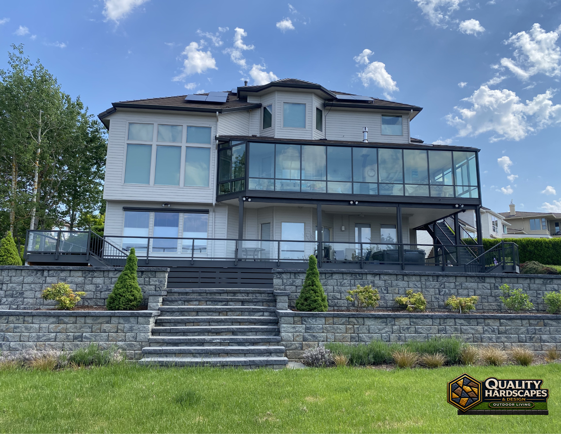 Large modern house with glass balcony, stone steps, and landscaped front yard under a blue sky