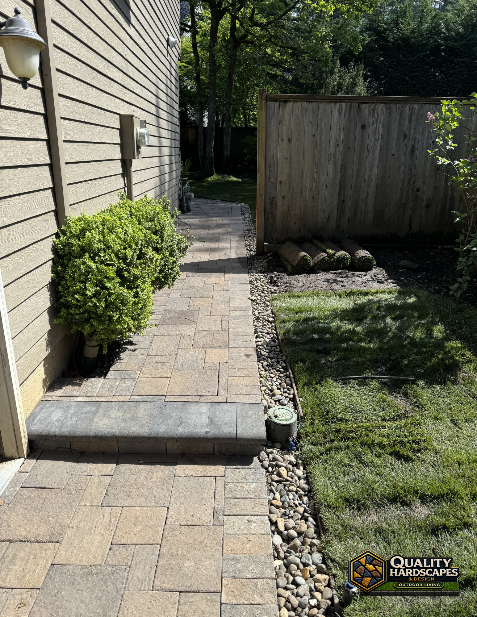Paver walkway beside house, with a small step, shrubs, and a wooden fence in a sunny yard.