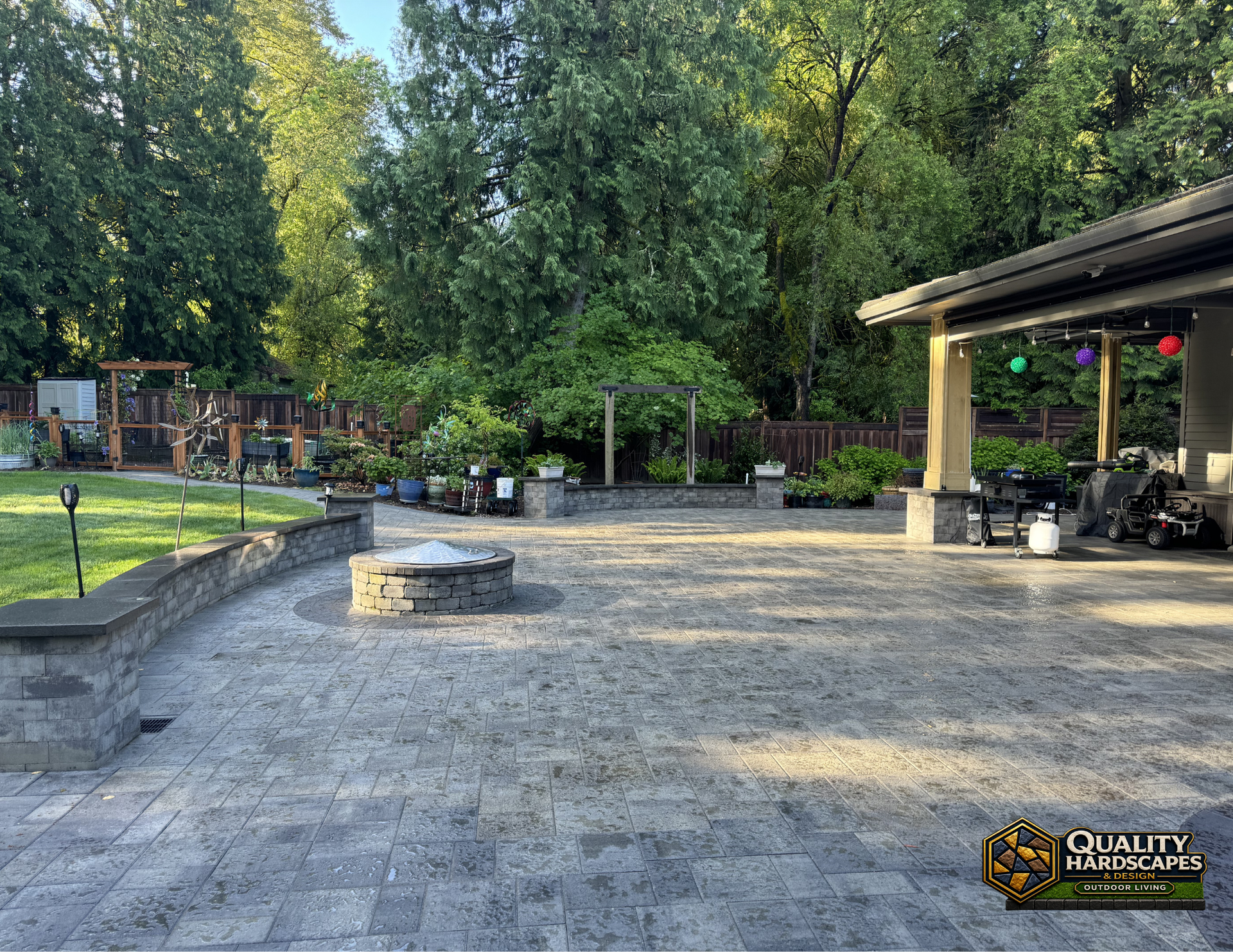 Stone patio with a circular fire pit, trees, and a covered pavilion in a landscaped backyard
