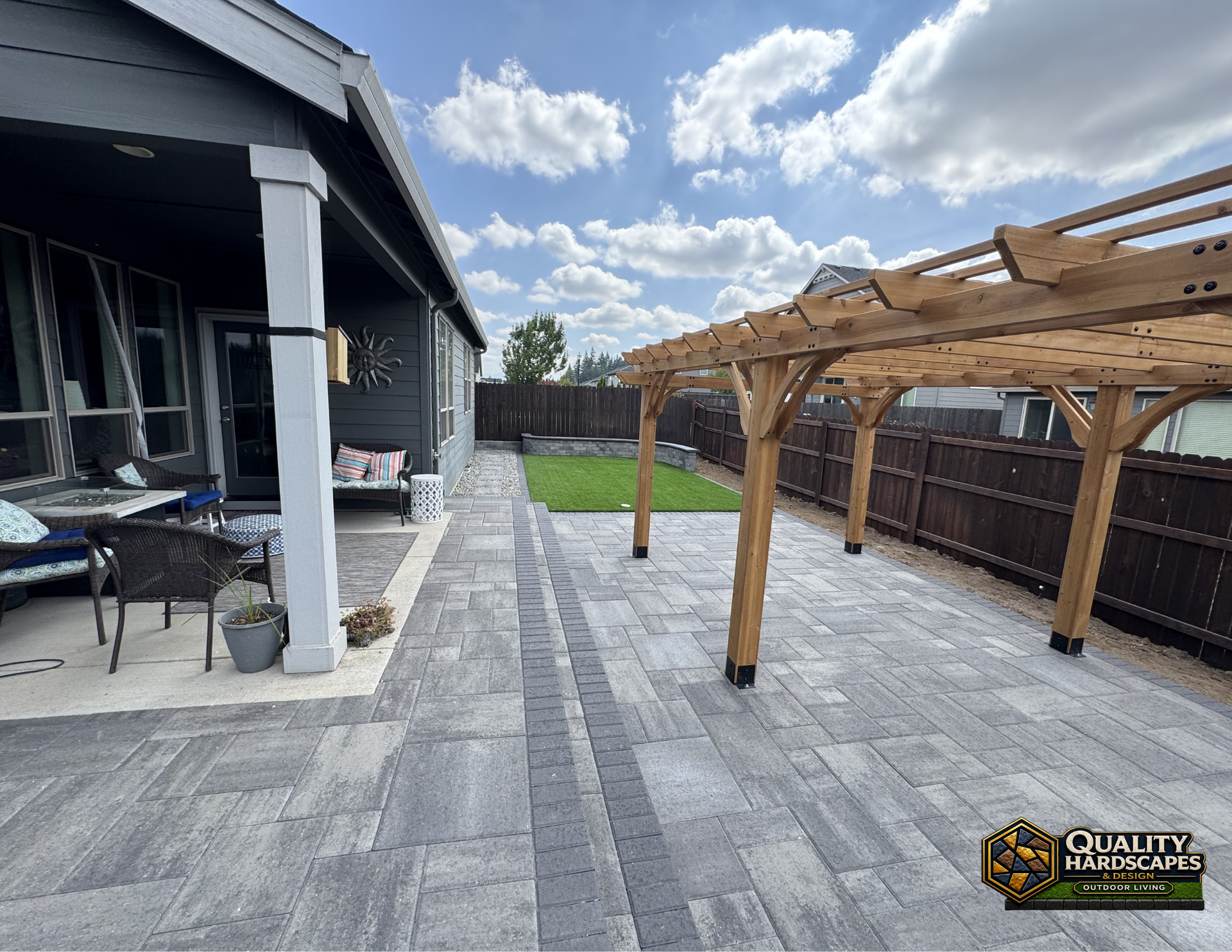 Backyard patio with gray pavers, covered seating area, and wooden pergola beside a house