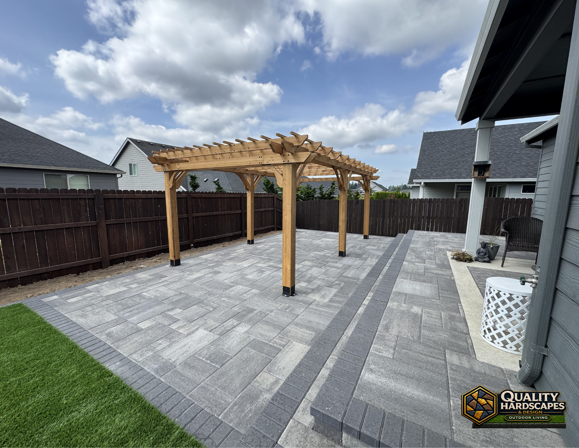 Backyard patio with stone pavers and a wooden pergola beside a lawn and fence under cloudy skies
