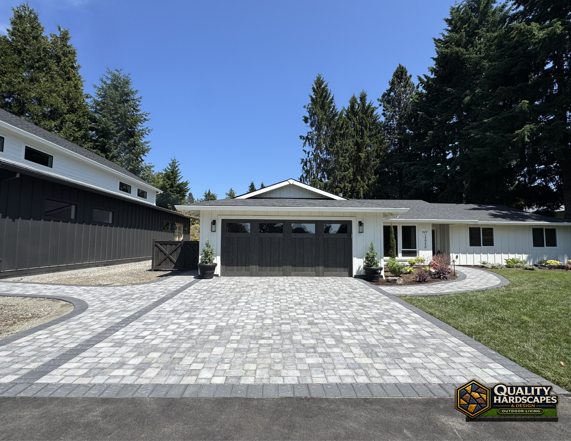 Single-story white house with gray garage door and paved driveway, framed by tall trees under a blue sky