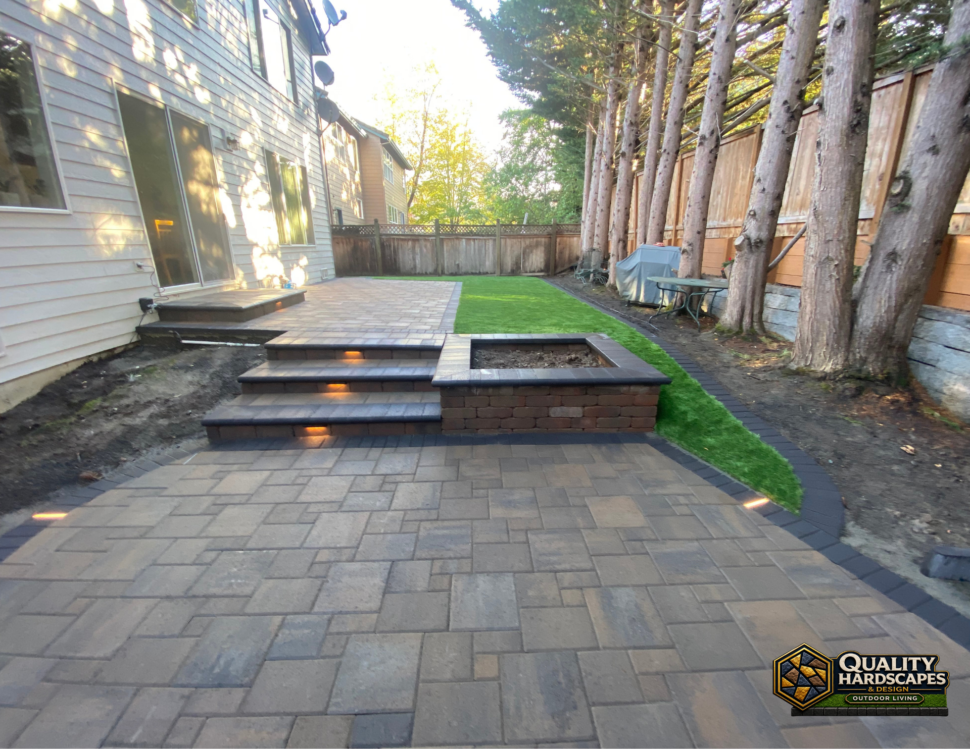 Backyard paver patio with wood steps, raised deck, and turf between a house and trees