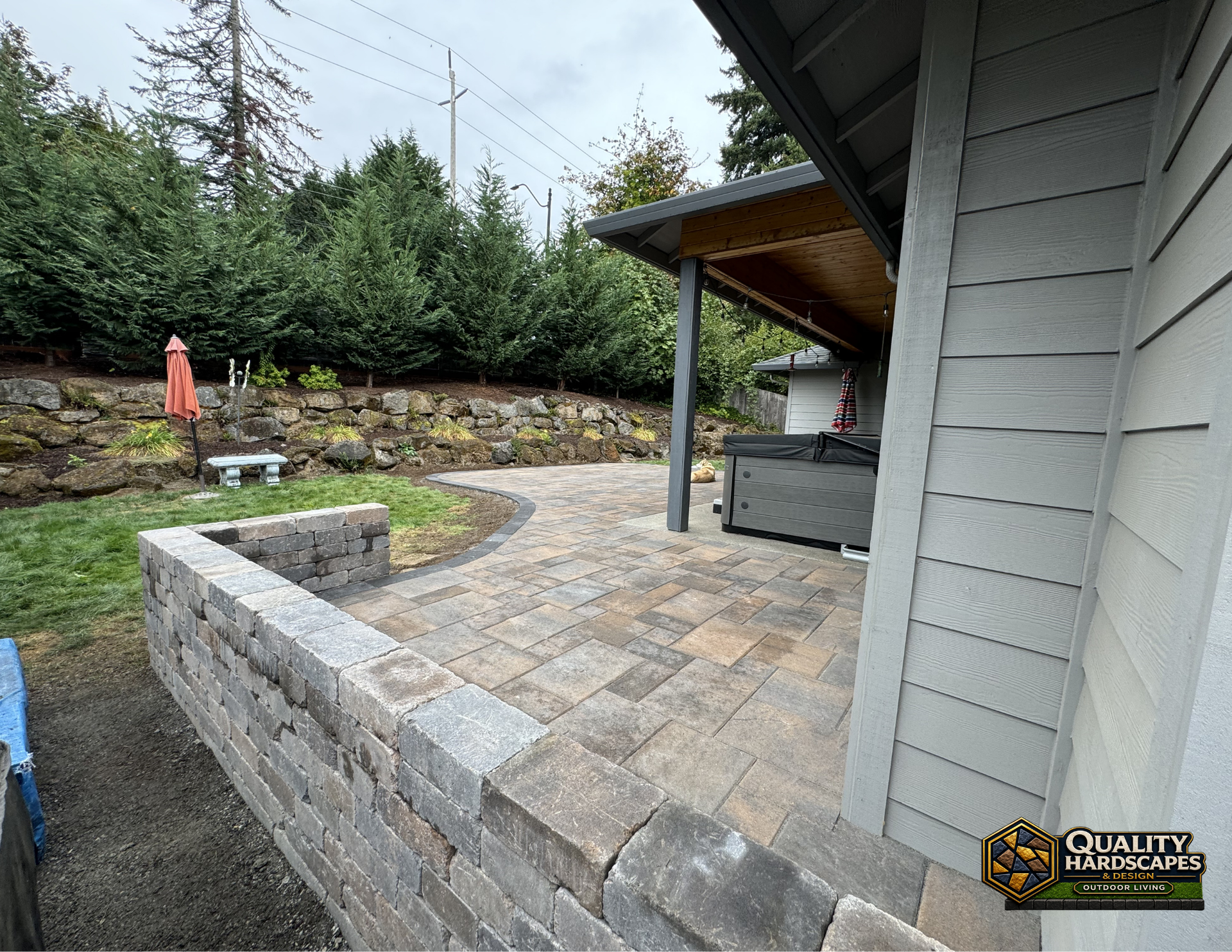 Stone patio beside house with covered porch, retaining wall, and landscaped yard beyond.