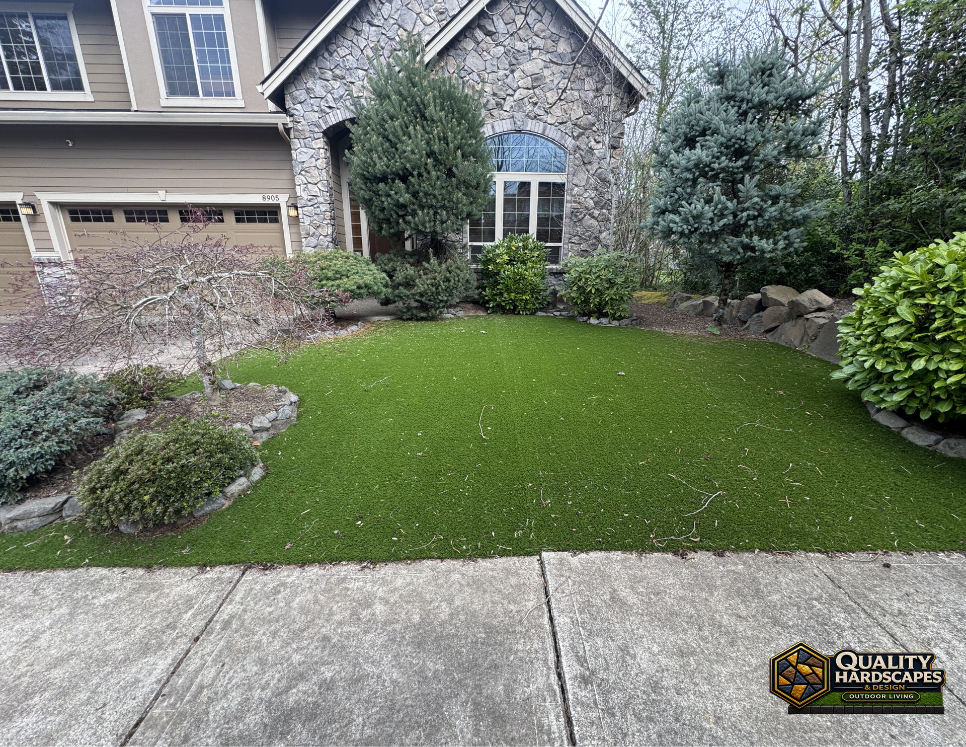 Front yard with lush green lawn, stone house, shrubs, and a concrete driveway or walkway in the foreground
